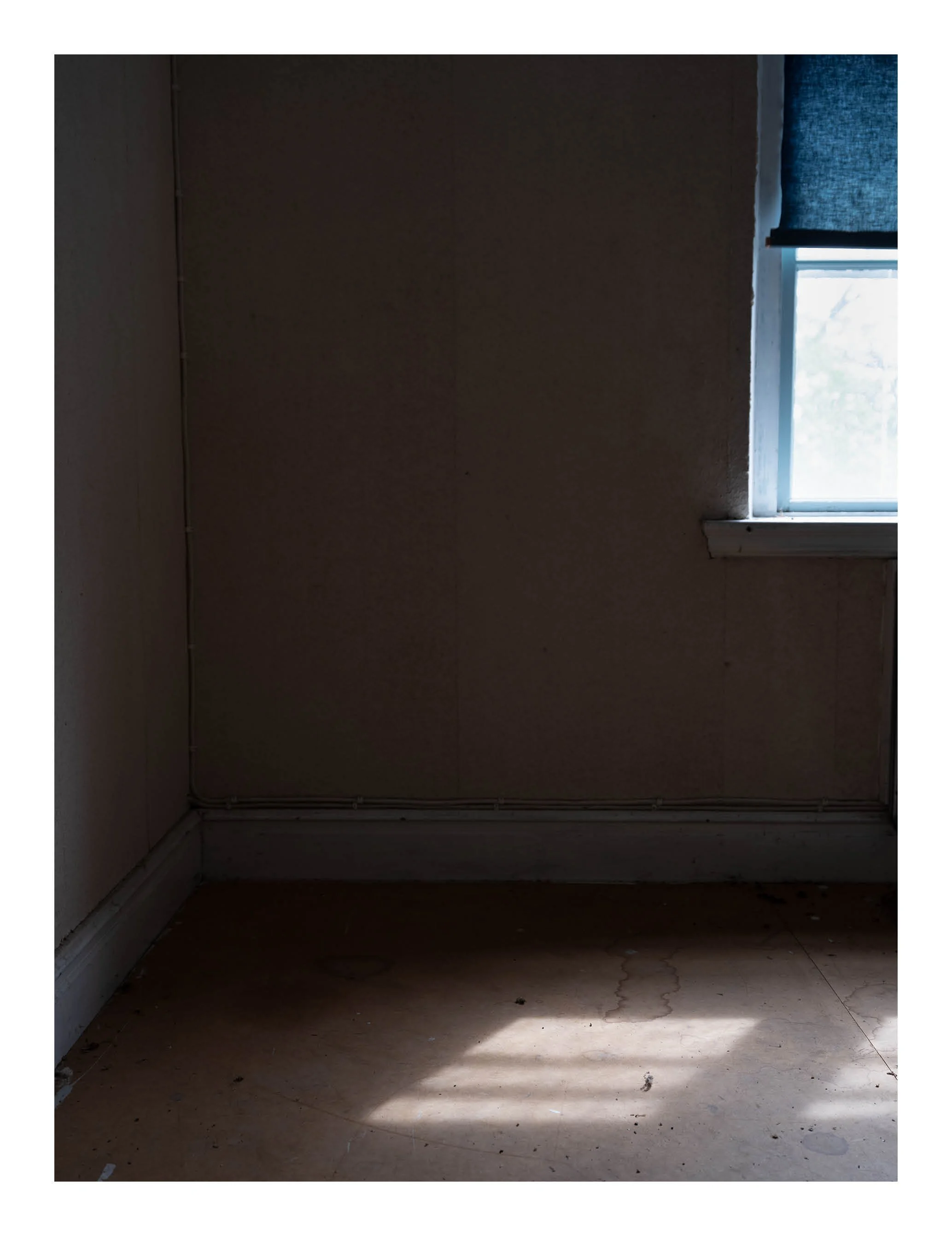 Empty room with a window and blue curtain, sunlight casting a shadow on the floor, beige walls, and a hardwood floor with some dirt and cracks.