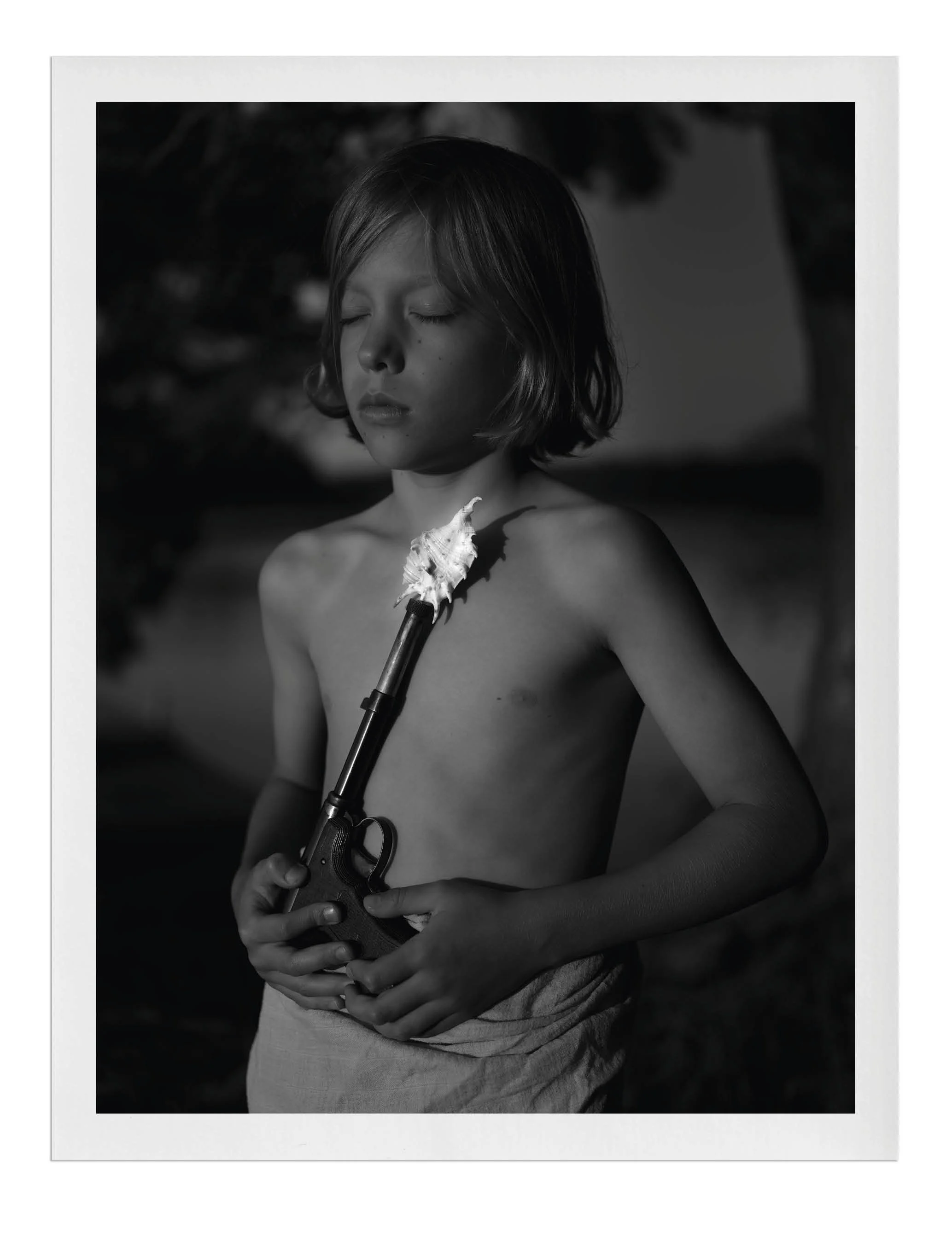 Black and white photo of a young boy with short hair, eyes closed, holding a toy revolver with a shell placed on his chest. The boy appears to be topless, with a neutral expression, and the background is blurred.
