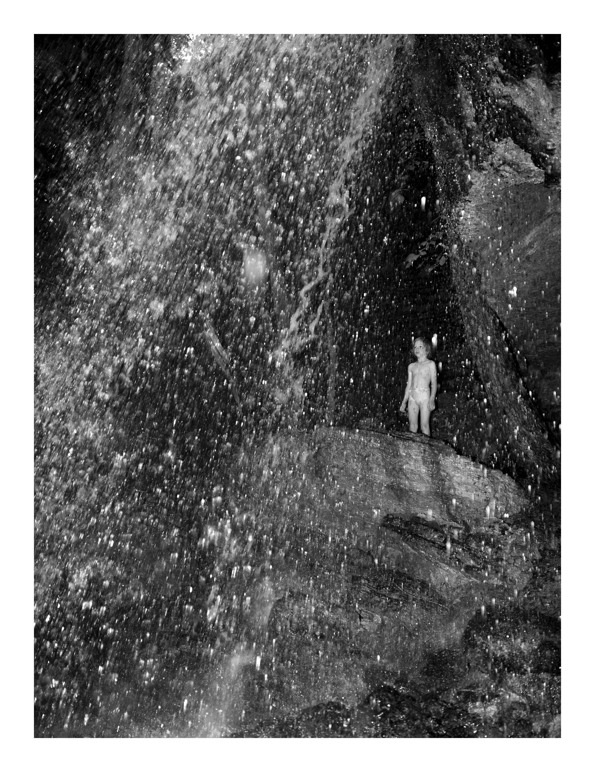 A boy standing under a waterfall with water splashing all around him in a dark environment.