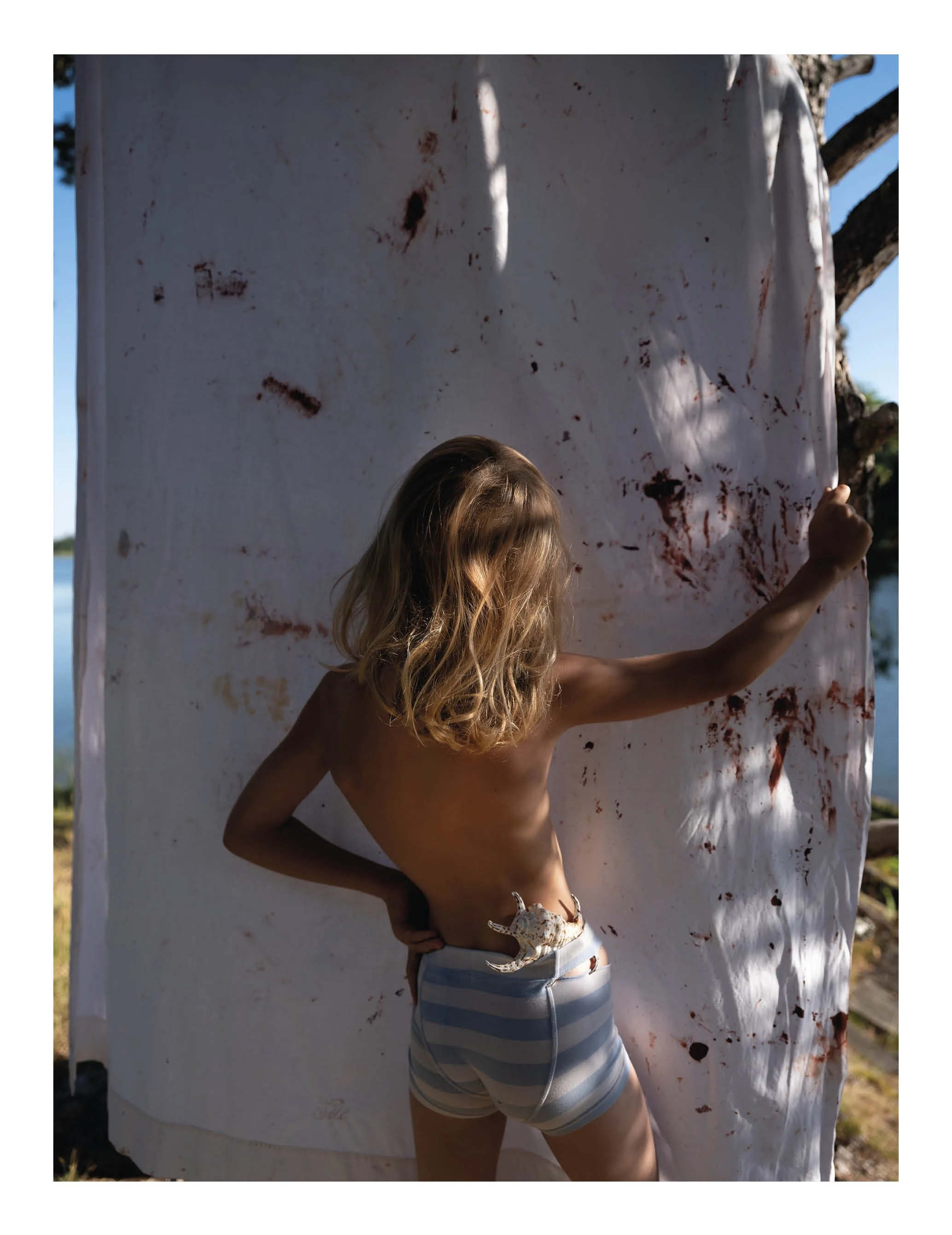 Person with long blonde hair wearing striped blue and white shorts, shirtless, standing in front of a white cloth with bloodstains, holding the cloth with one hand, outdoor setting with trees and blue sky.