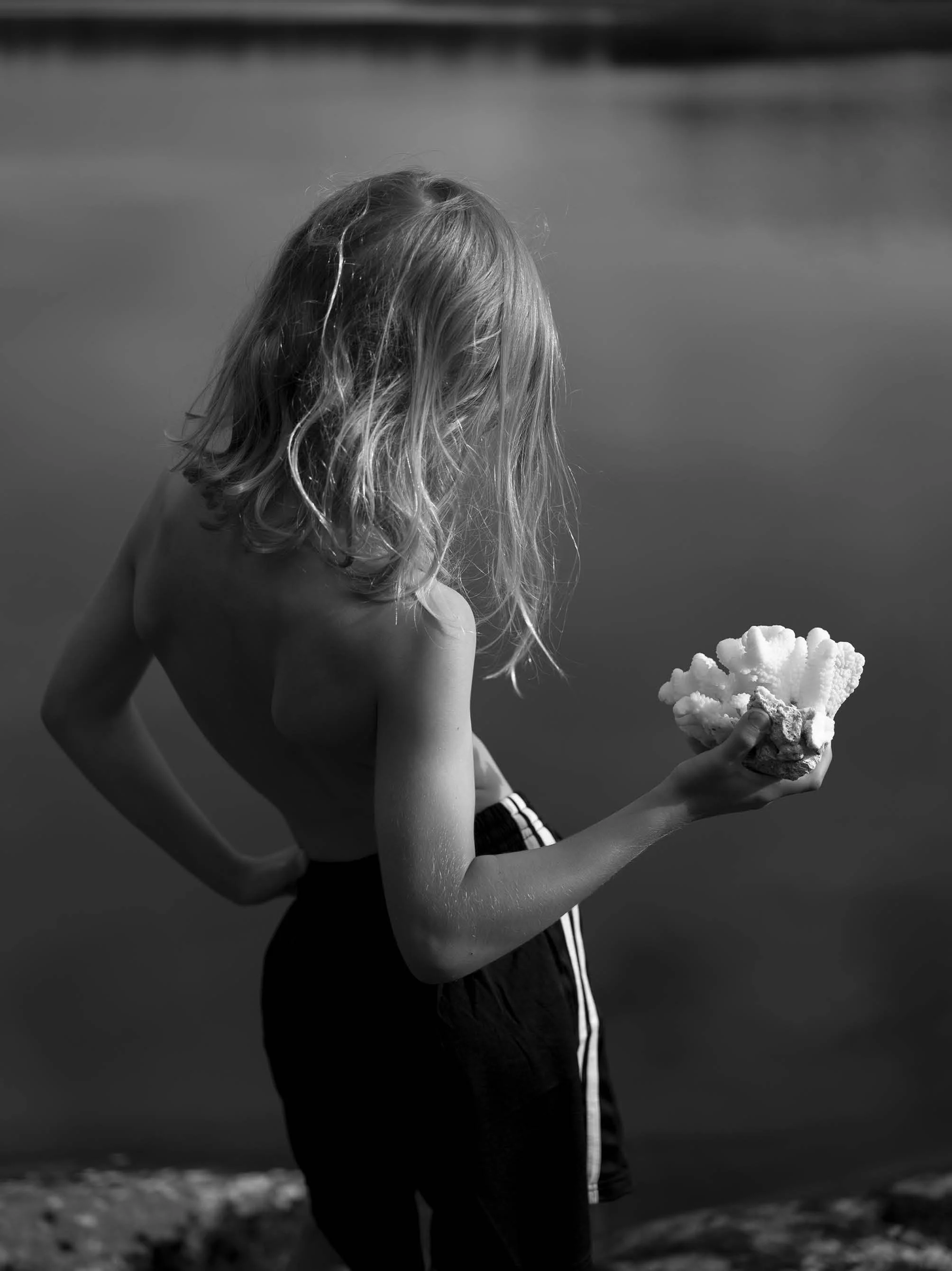 A black-and-white photo of a young girl with long, curly hair standing outdoors near the water, holding a piece of coral in her hand, with her head slightly bowed.