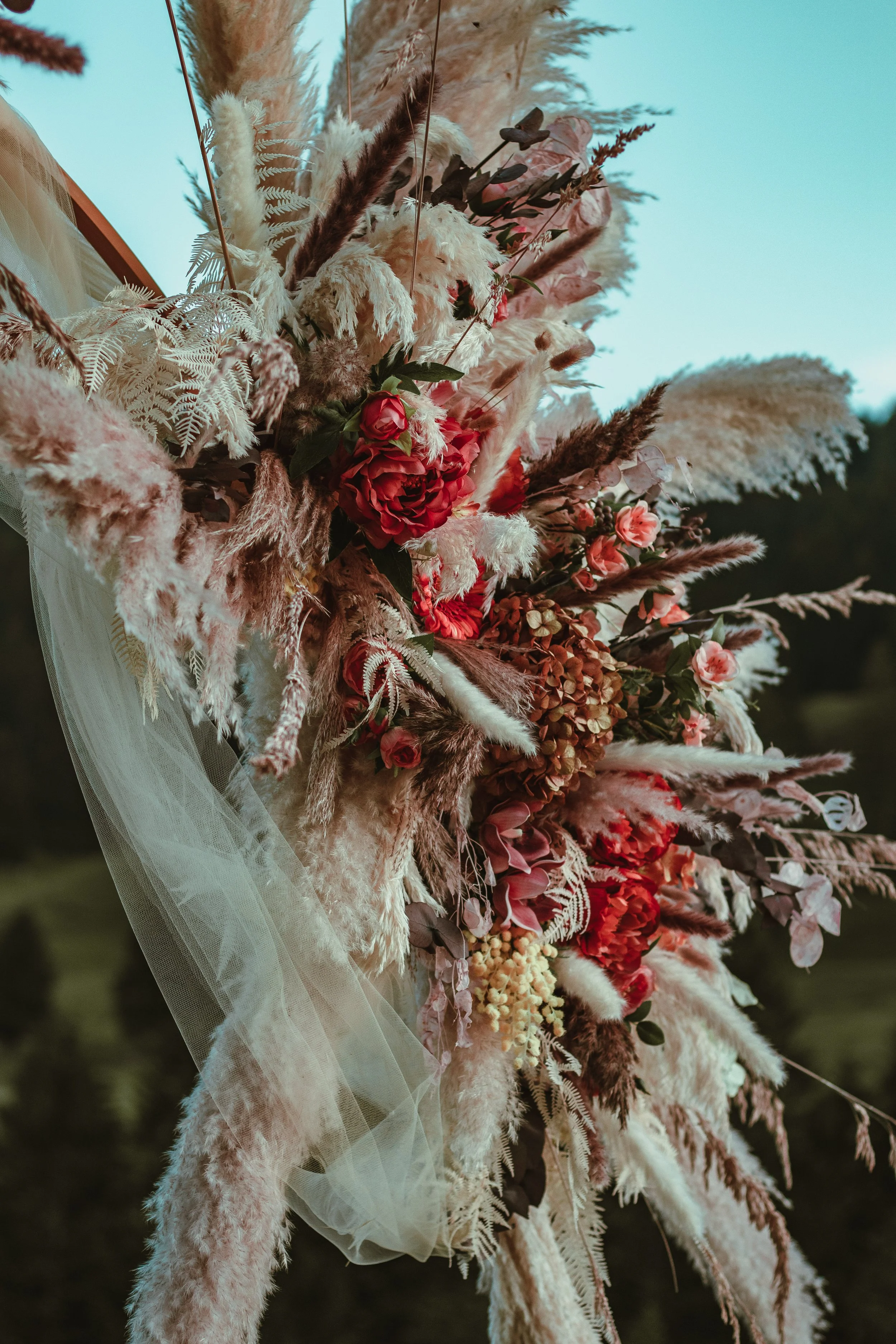 A large floral arrangement with dried and fresh flowers, including pampas grass, roses, and various greenery, wrapped in white tulle fabric with a mountain in the background- complete setup for a romantic proposal in Switzerland, Rhine Valley