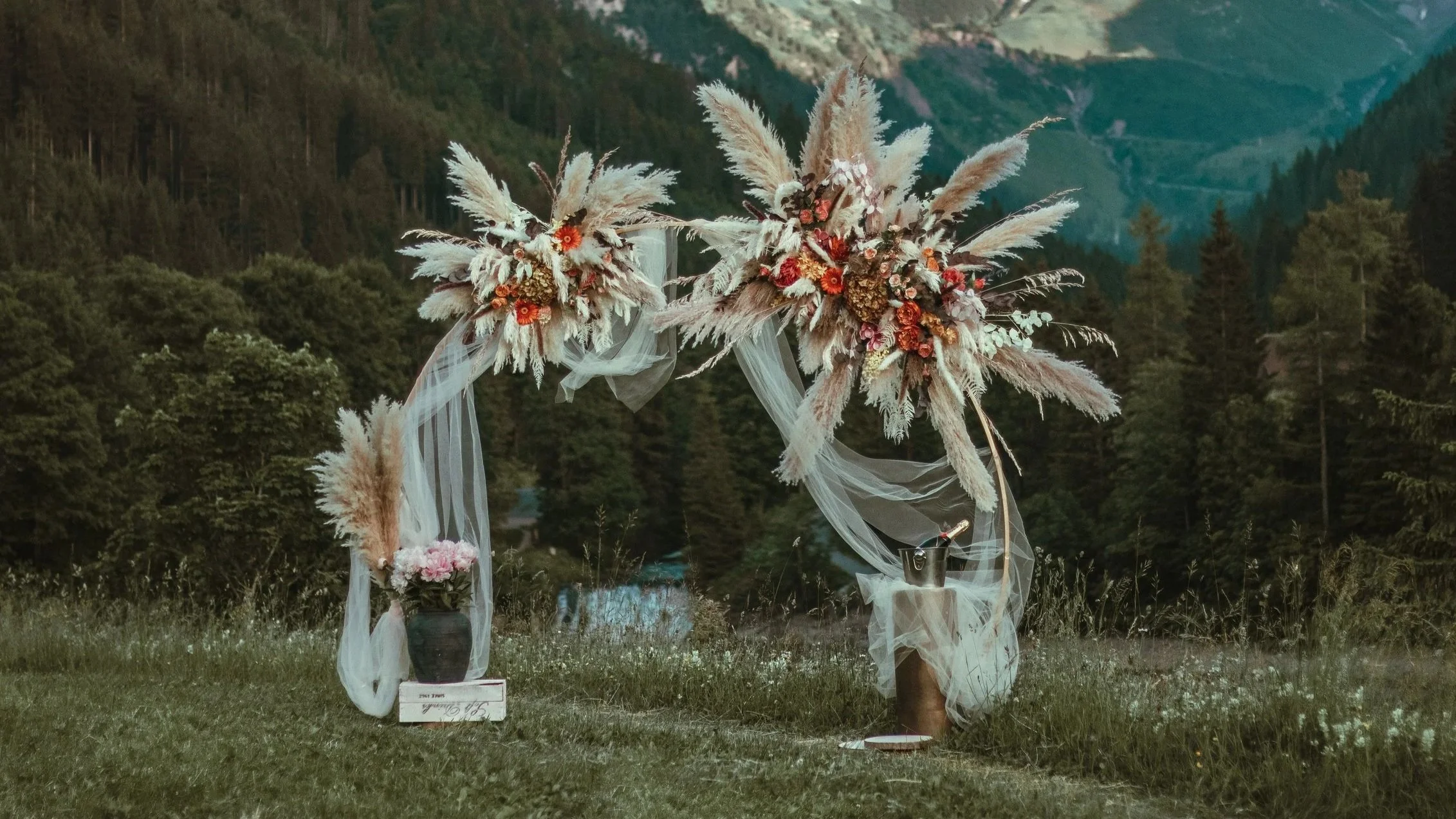 An outdoor proposal setup with a floral arch decorated with pampas grass and pink and red flowers, set against a scenic mountain and forest background in the Swiss Alps for a romantic and effortless engagement photoshoot
