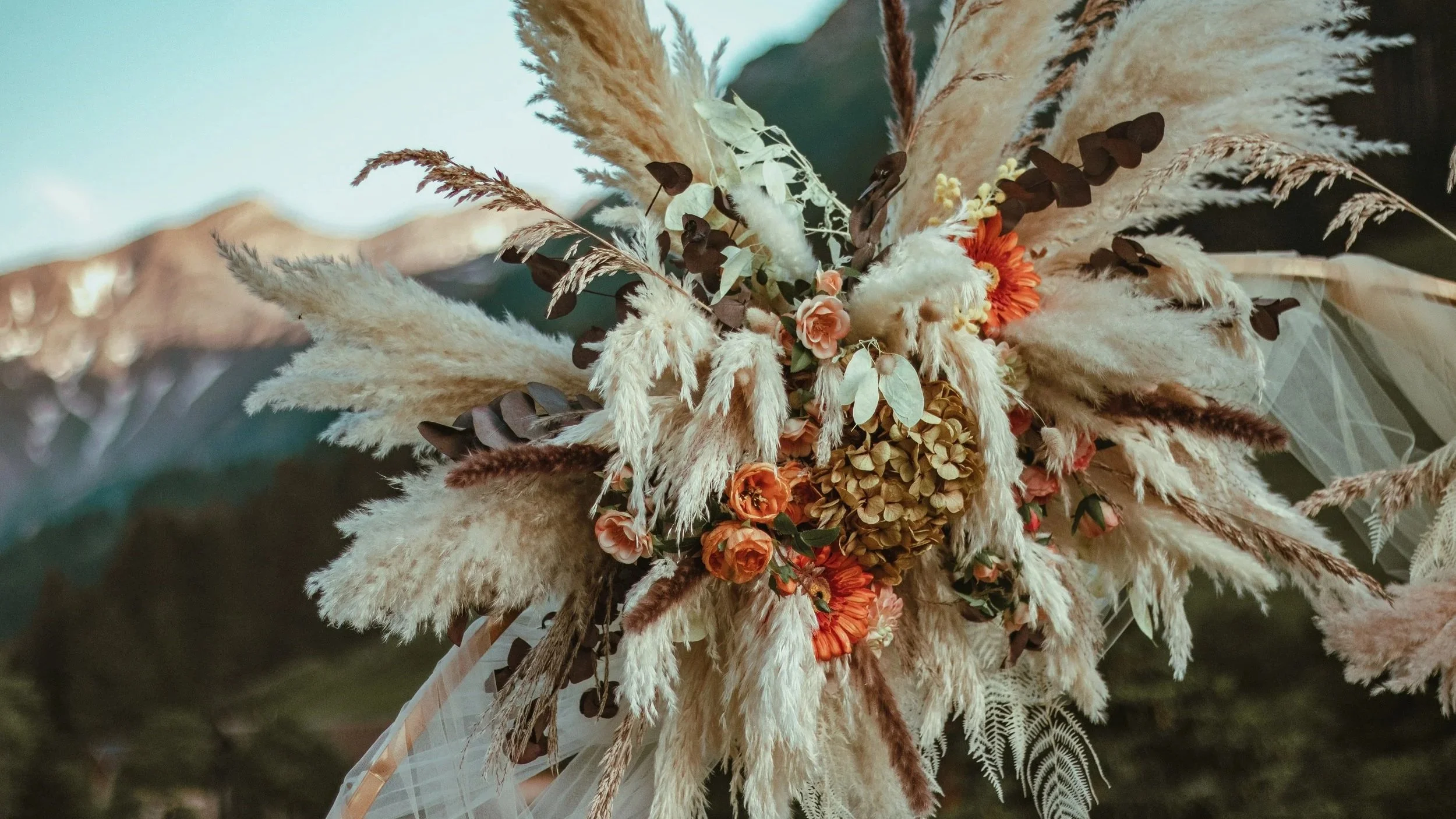 A floral bouquet with dried grasses, faux flowers, and greenery, held with a white ribbon and some sheer fabric- romantic and complete setup for a surprise proposal in Switzerland, with or without a photoshoot and video of the proposal moment