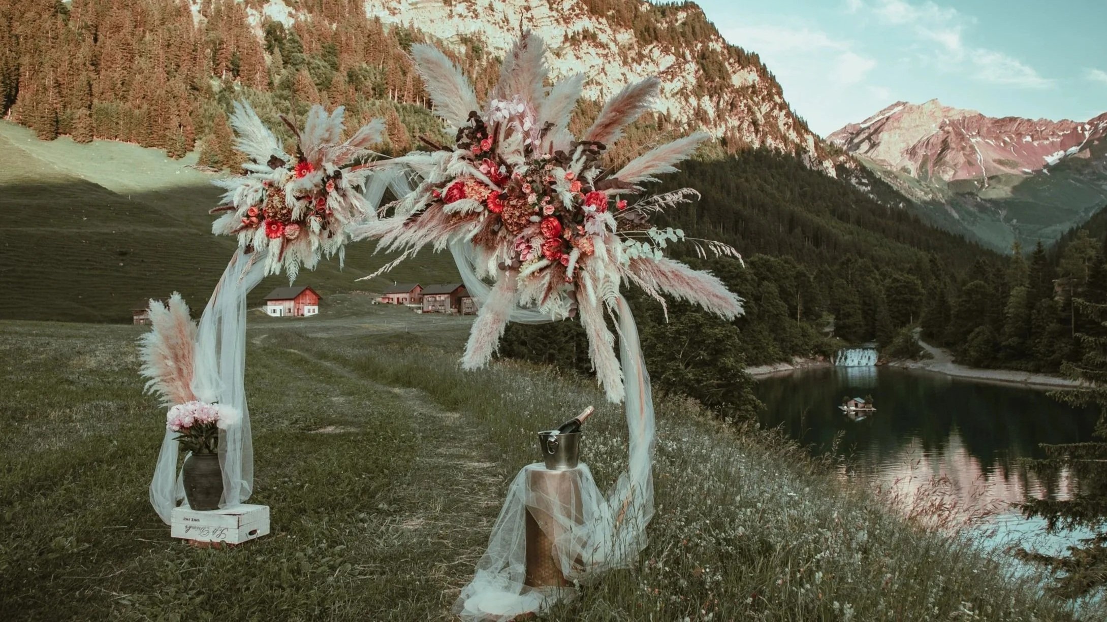 A floral arch decoration for a surprise proposal set outdoors near a lake with mountains and pine trees in the background- complete setup for a beautiful and effortless engagement 