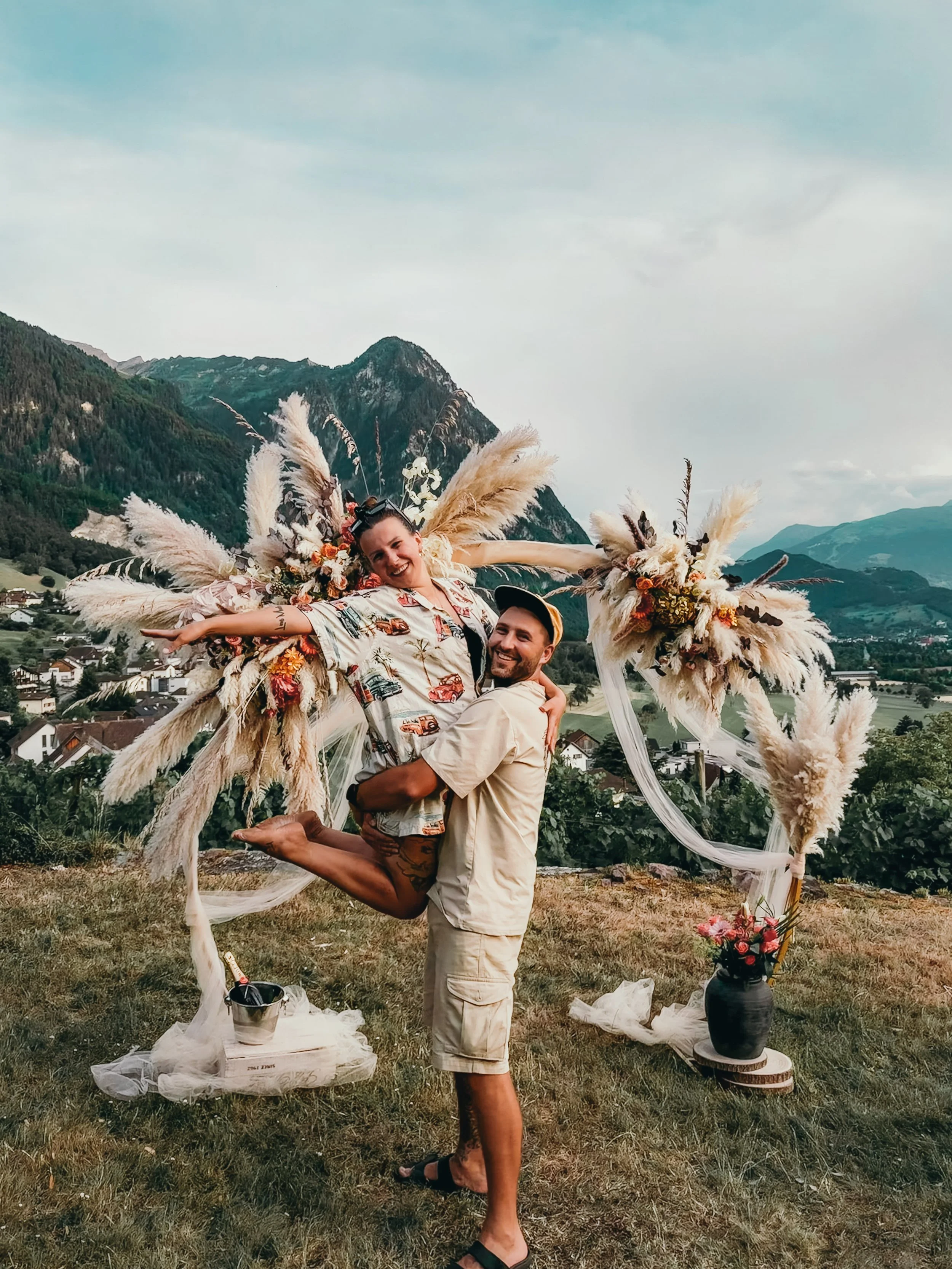 A couple celebrating with a large, decorative floral and pampas grass arch, a romantic setup in an outdoor scene with mountains in the background. Happy couple in a proposal photoshoot- very romantic and beautiful