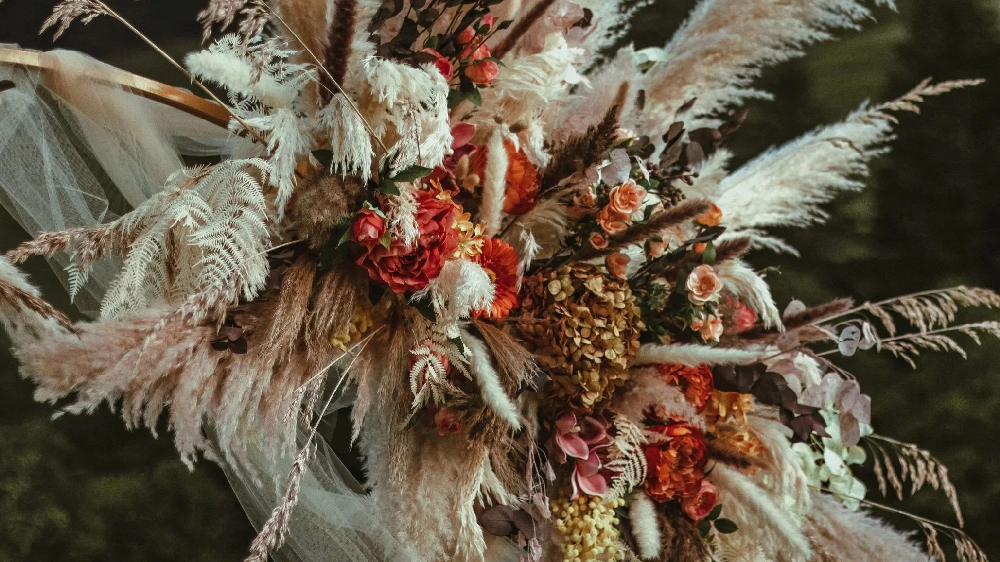 A close-up of a bouquet with various dried flowers and grasses in shades of white, pink, orange, and brown, arranged with greenery and decorative tulle fabric- acomplete romantic setup for an engagement in the Swiss Alps, near lakes and mountains.