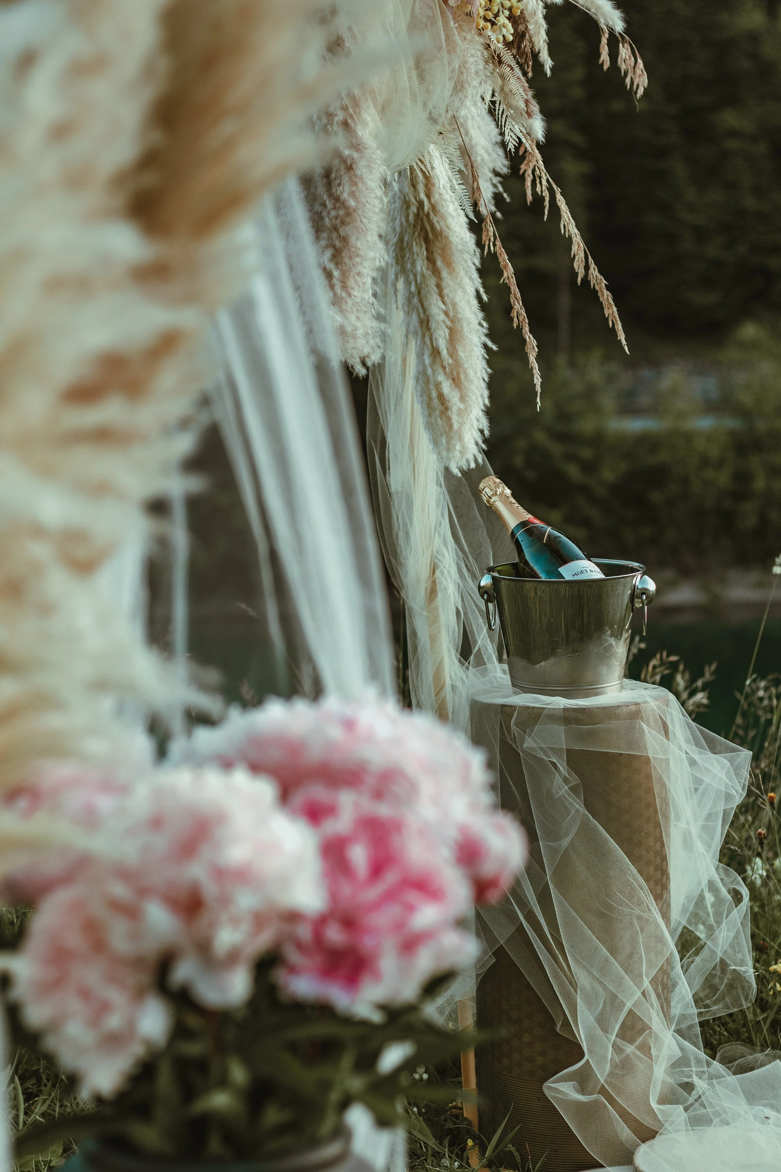 A bouquet of flowers in the foreground, out of focus, with a table arrangement in the background and Moët champagne chilling in an ice bucket, adorned with sheer, flowing fabric- romantic setup for a scenic outdoor proposal in Switzerland