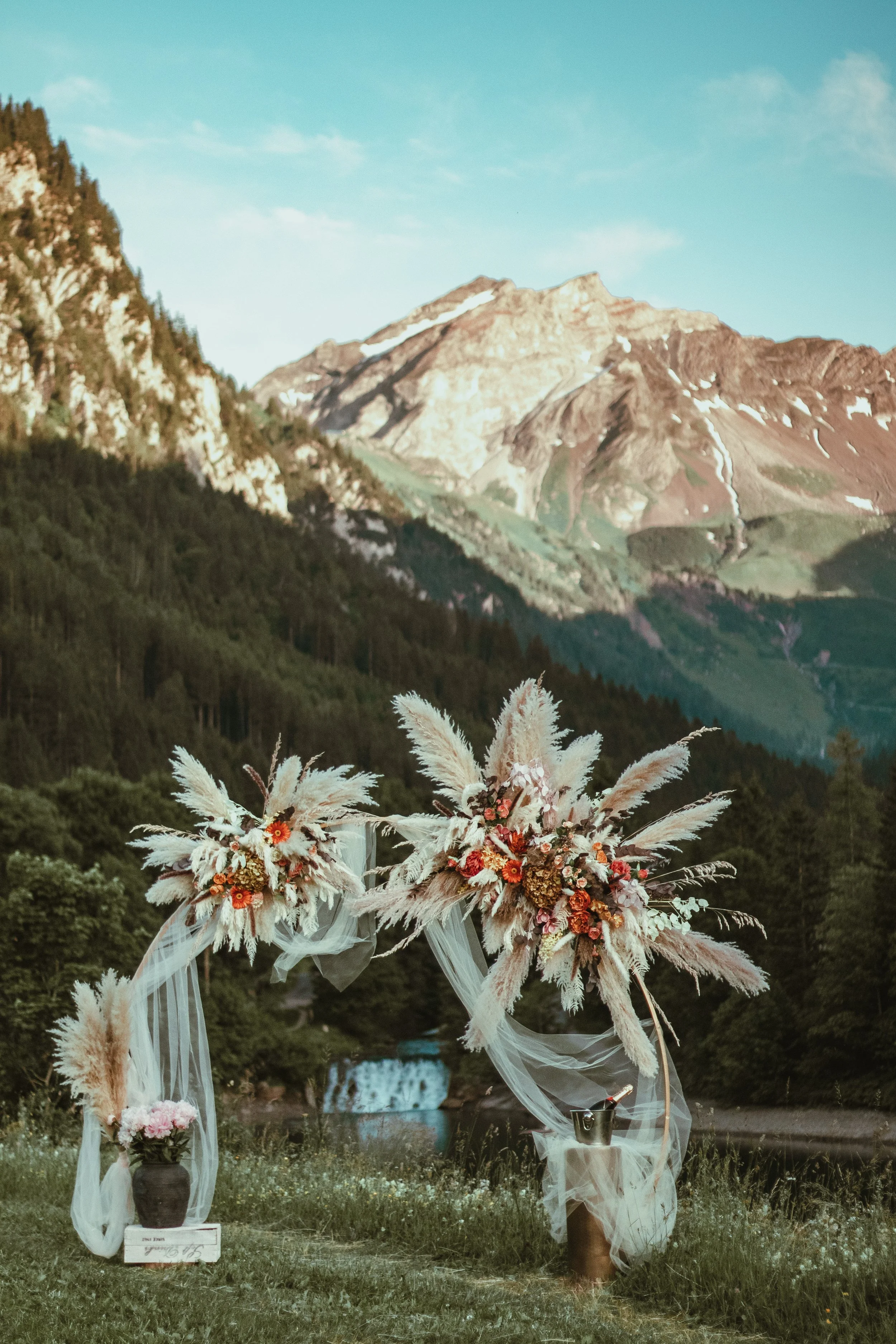 Outdoor wedding altar decorated with floral arrangements, pampas grass, and flowing fabric, set against a mountain-and-forest backdrop. Romantic setup for a proposal in a scenic outdoor area for a surprise proposal photoshoot in the Swiss Alps. 