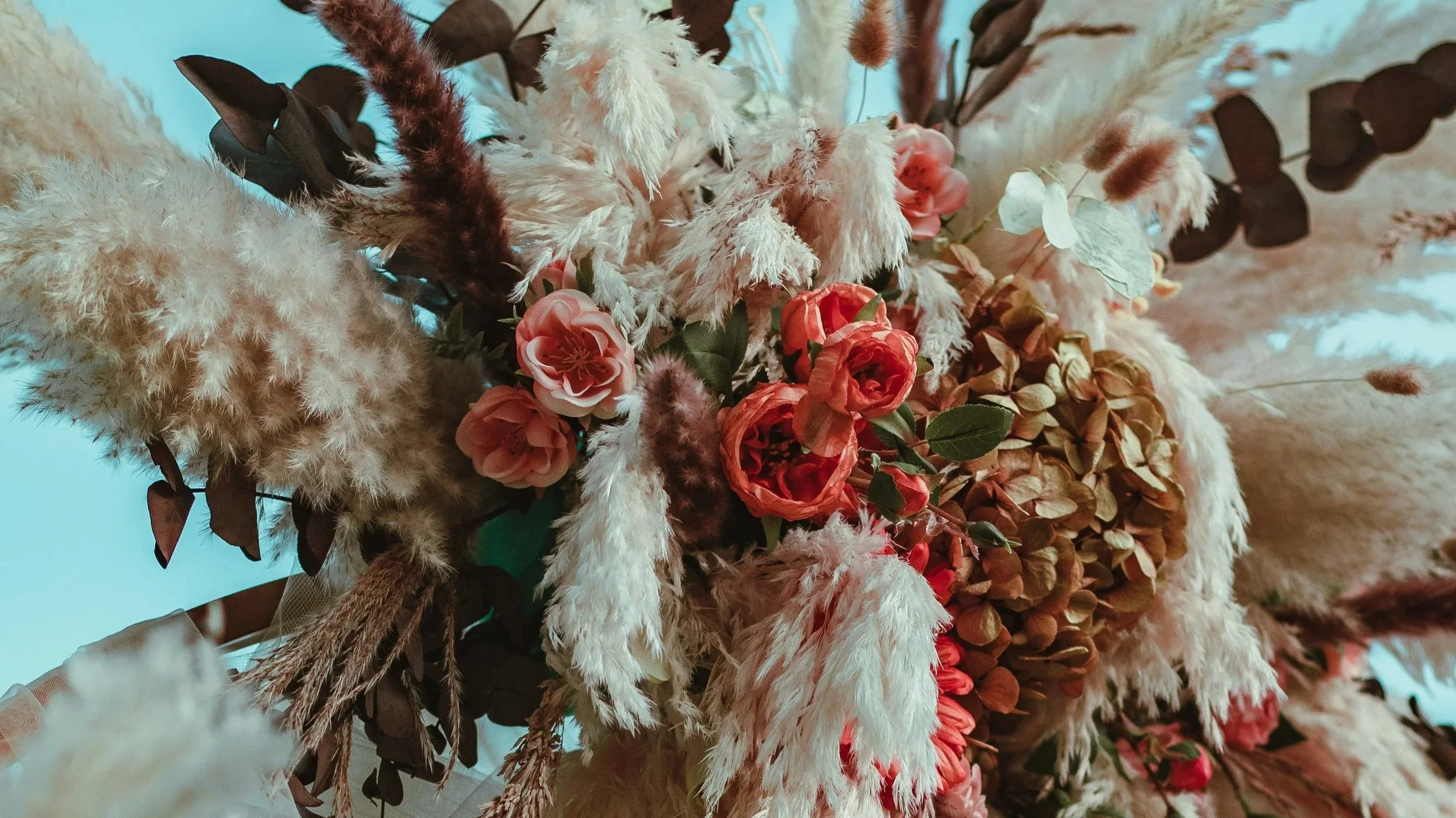A bouquet of dried flowers, including pink roses, white fluffy plumes, brown leaves, and other dried botanical elements on an arch, romantically decorated for a surprise proposal photoshoot with a mountain backdrop in Switzelrand and Liechtenstein