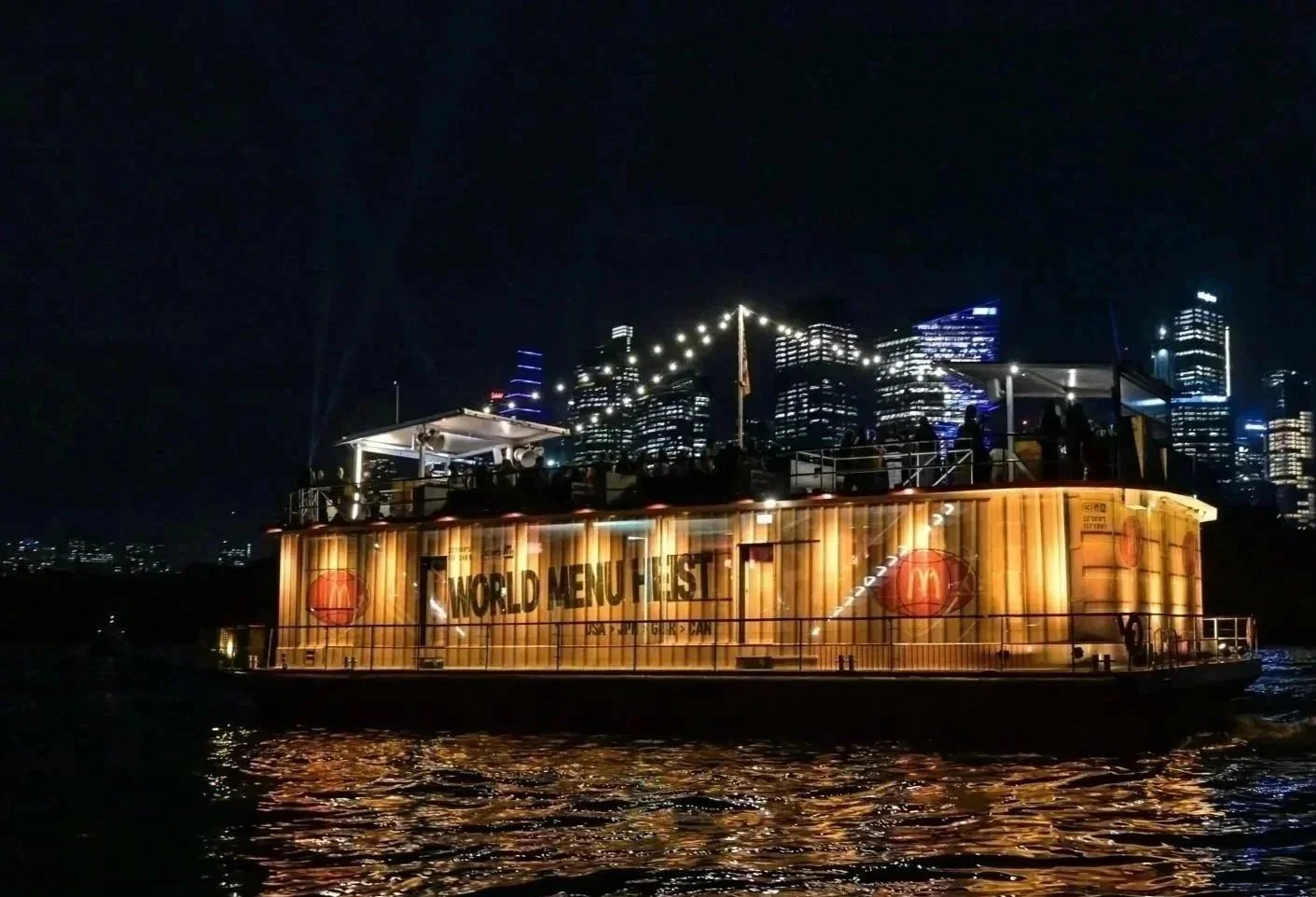 Illuminated McDonald's Menu Heist activation floating restaurant boat on nighttime water with a city skyline in the background. Glass island boat wrap with print and one way vision in sydney harbour.