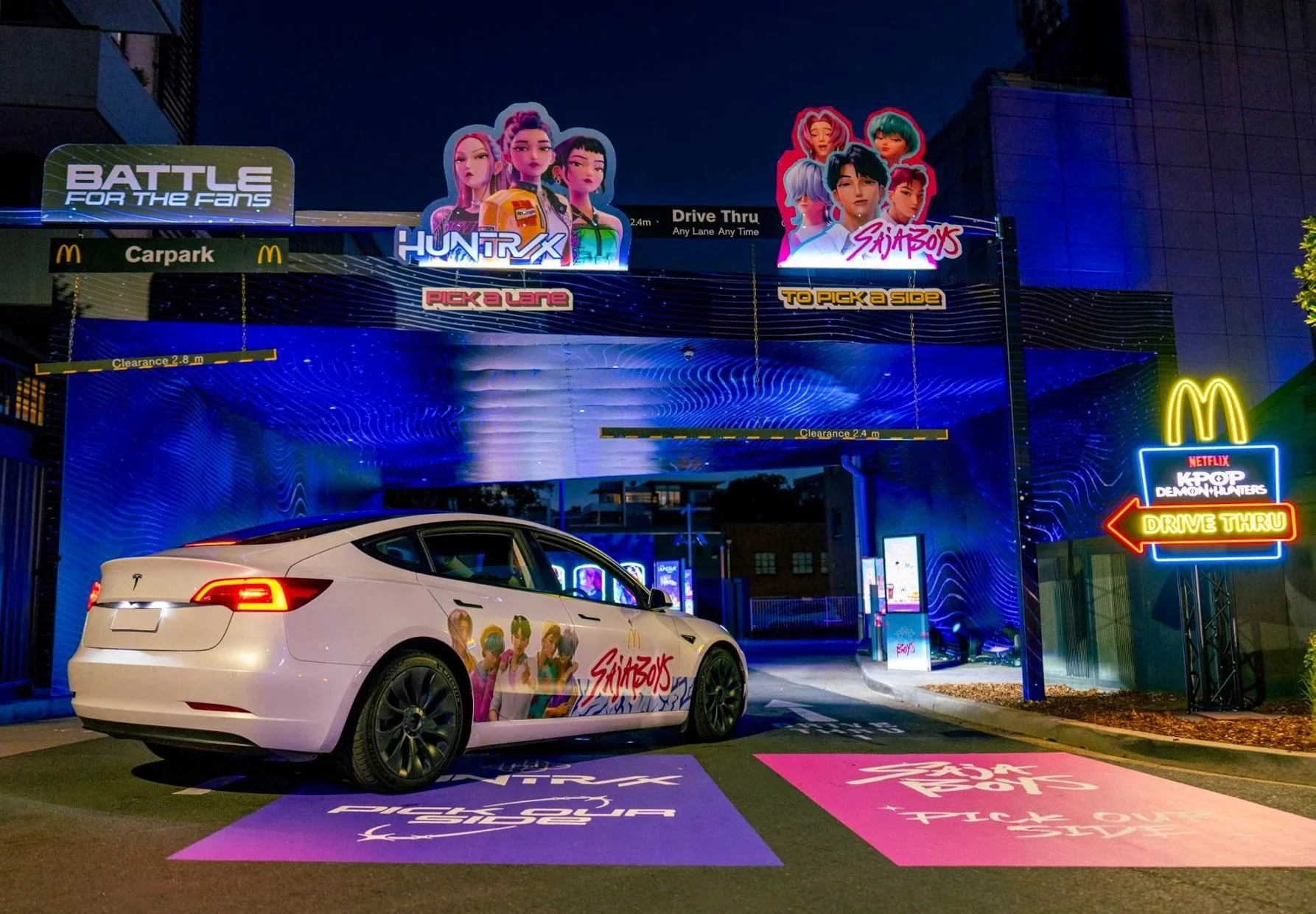 Nighttime McDonald’s drive-thru activation decorated for a K-pop demon hunters promotion, featuring illuminated HUNTRIX and Saja Boys signs and a branded white Tesla. Floor graphics, car decals and graphics, acp panel with print, lightbox prints.