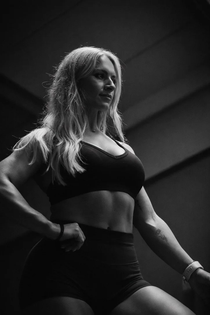 A black and white photo of a woman with long, wavy hair wearing a sports bra and high-waisted shorts, looking confident with her hand on her hip.