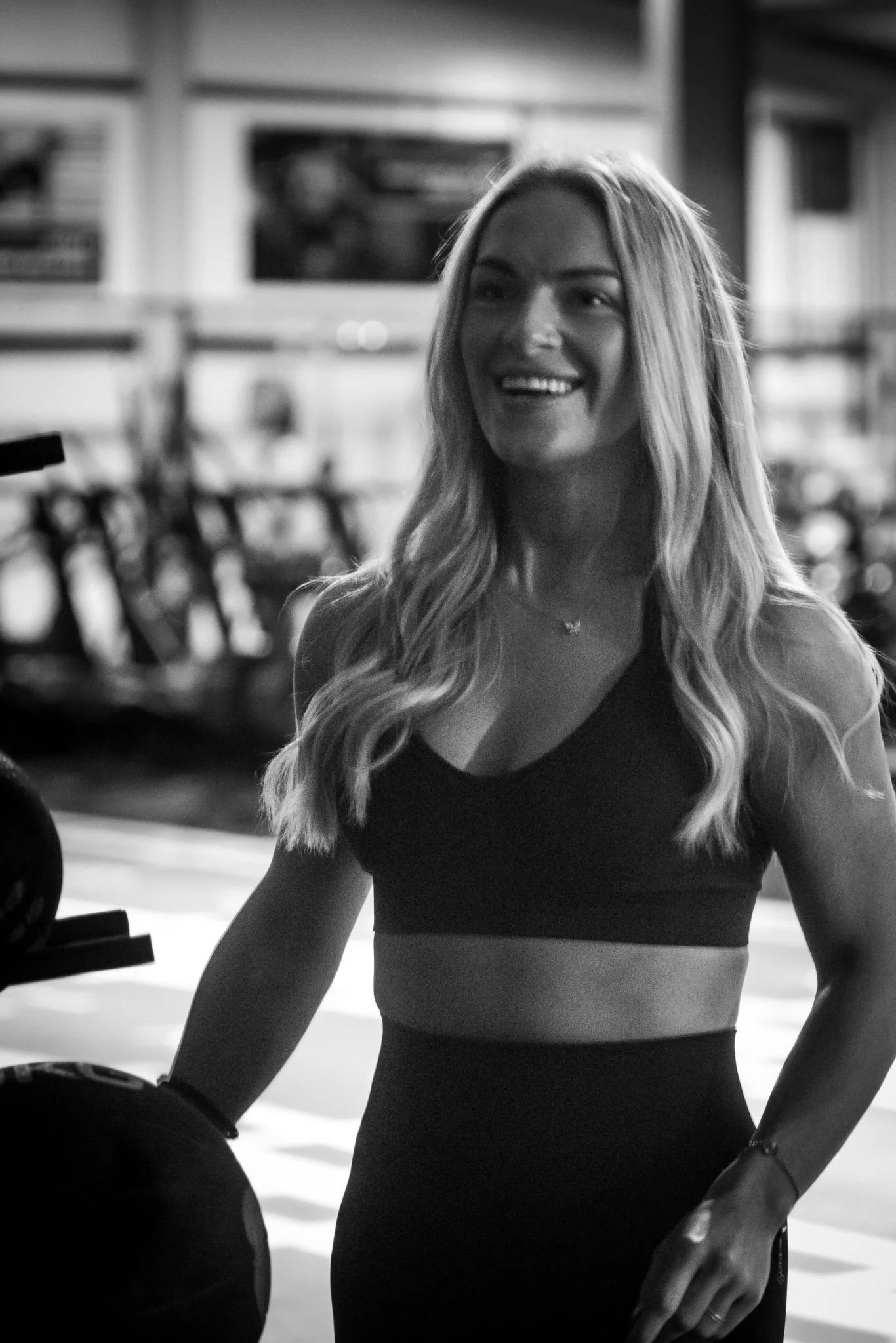 A woman with long wavy hair smiling in a gym setting, wearing workout attire, with gym equipment visible in the background.