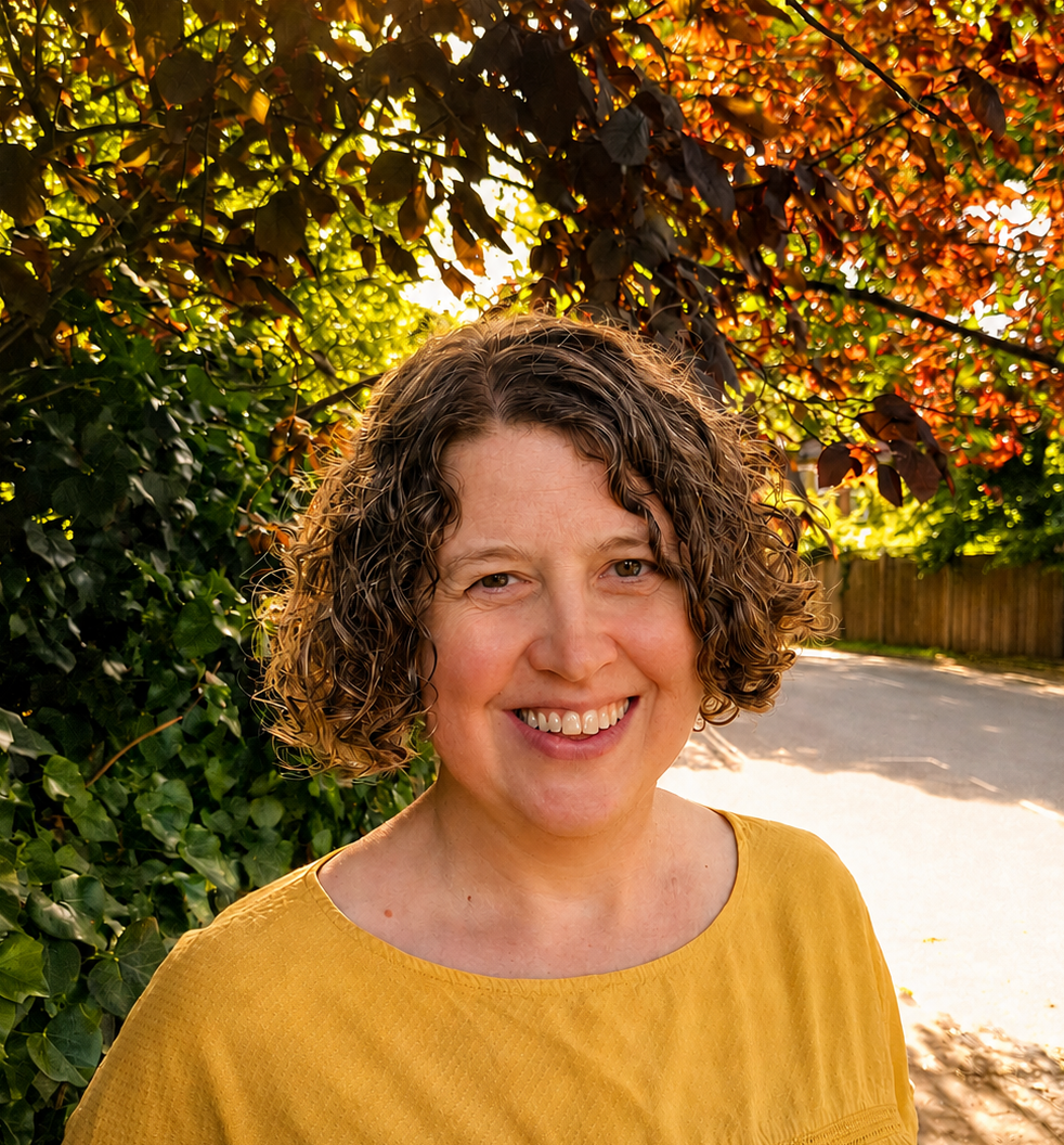 Photo of a woman with dark curly hair, wearing a yellow round necked top.  There's green and red foliage and a road in the background.
