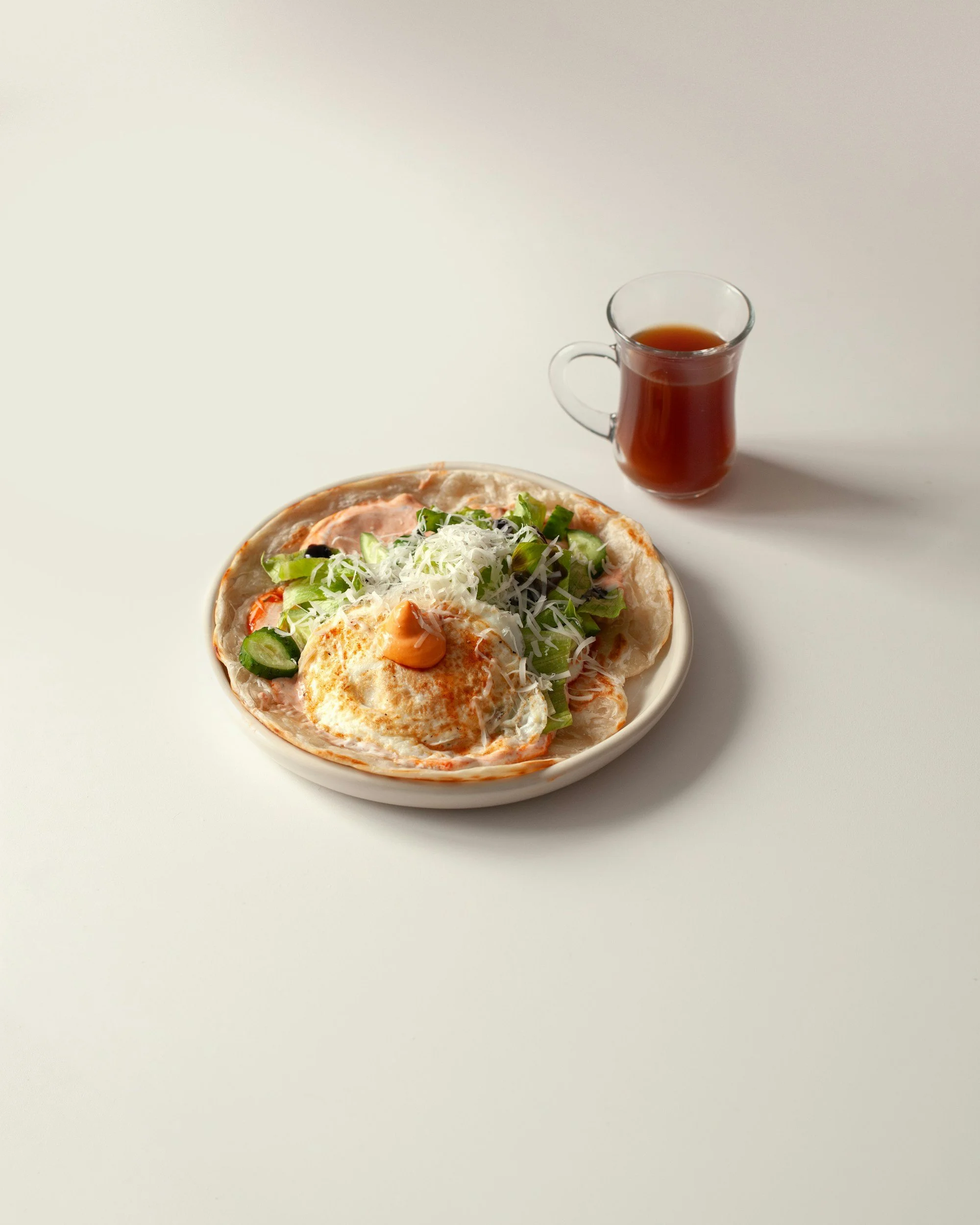 A pizza topped with cheese, vegetables, and an egg, on a white plate, with a glass of dark tea nearby, against a white background.