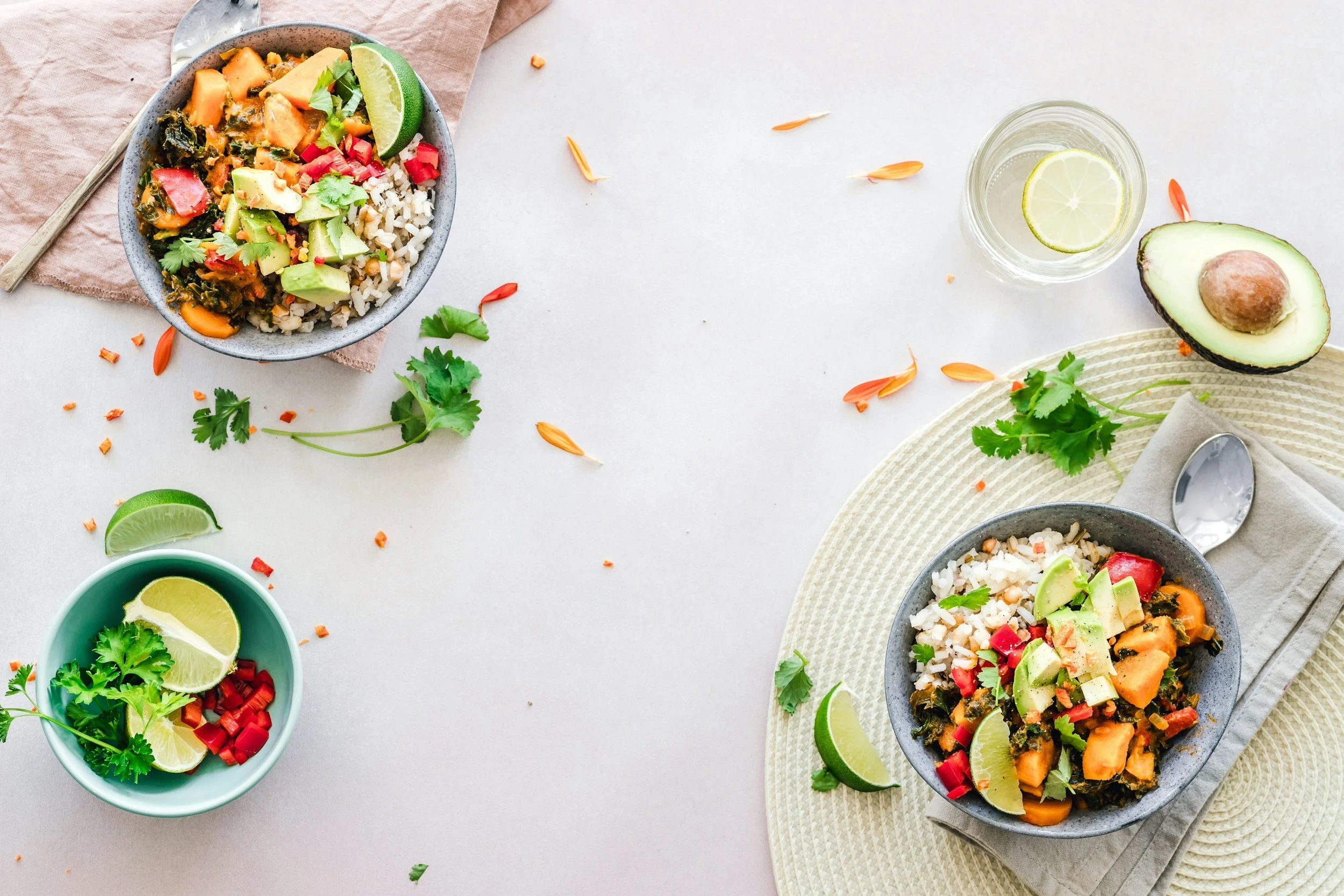 Bowls of rice and vegetable curry with lime wedges, cilantro, and avocado, surrounded by smaller bowls of chopped red peppers, lime slices, and cilantro, on a white surface with scattered petals and leaves.