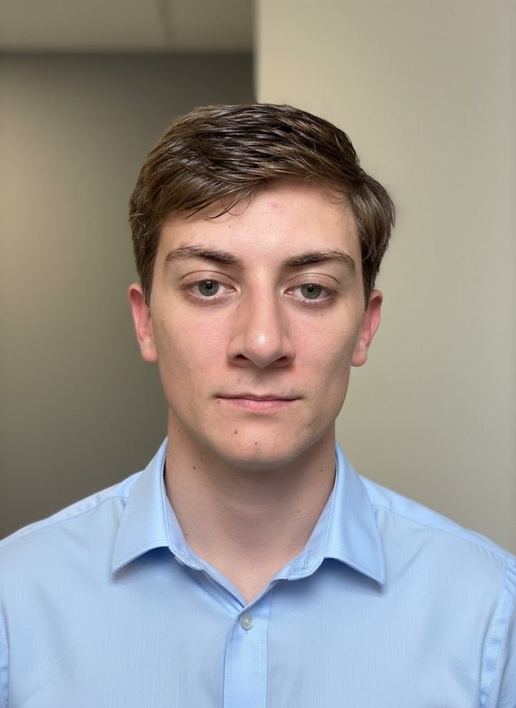 A young man with short brown hair and blue eyes, wearing a light blue collared shirt, looking directly at the camera in an indoor setting.
