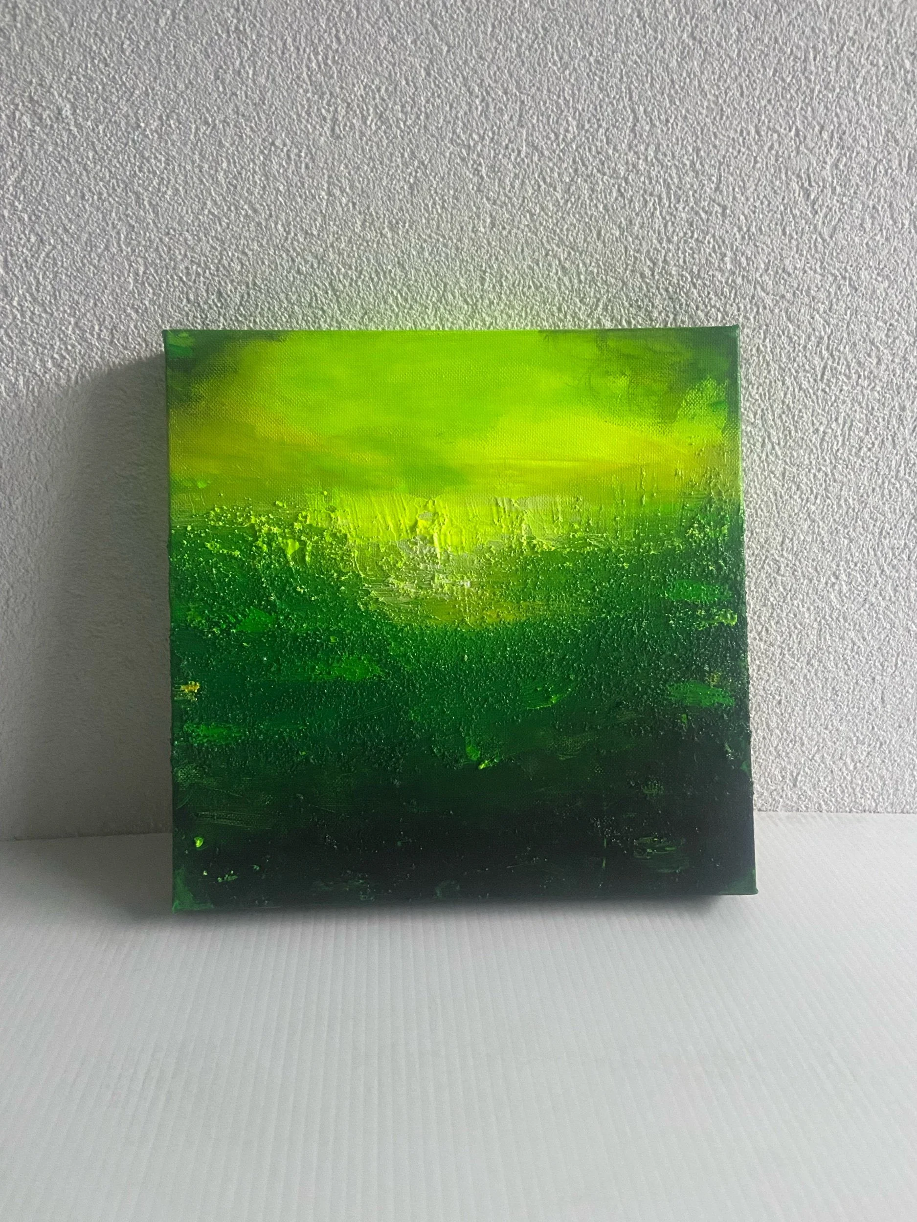 An abstract painting with shades of green, yellow, and black, displayed on a white textured wall.