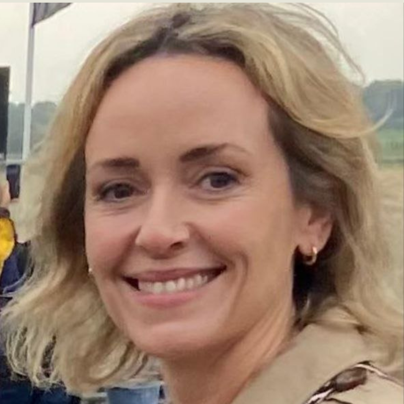 Close-up of a smiling woman with blonde hair and hoop earrings, outdoors in a rural area.