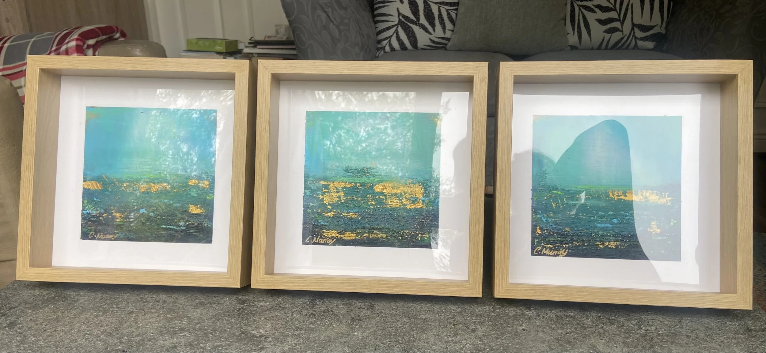 Three framed paintings of ocean scenes with blue, green, and gold hues, leaning against a surface indoors.