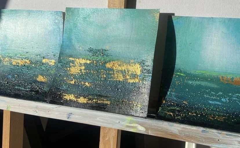 Three abstract landscape paintings on display, featuring textured scenes with blue, green, yellow, and black colors.