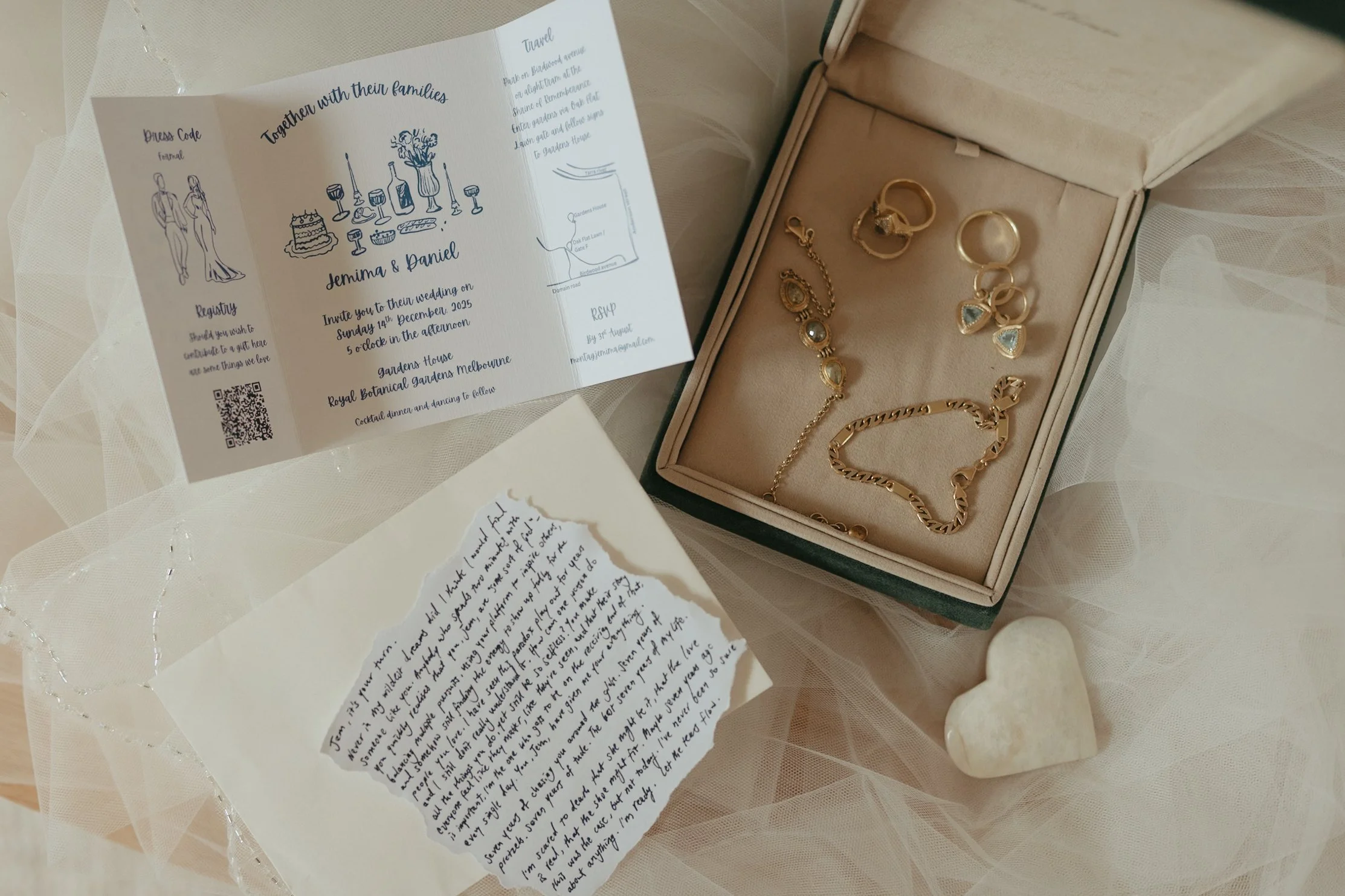 Open jewelry box containing gold rings, earrings, and bracelets on a beige cushion. Next to it, wedding invitation with illustrations of couples, flowers, place, and date details, and a handwritten note.