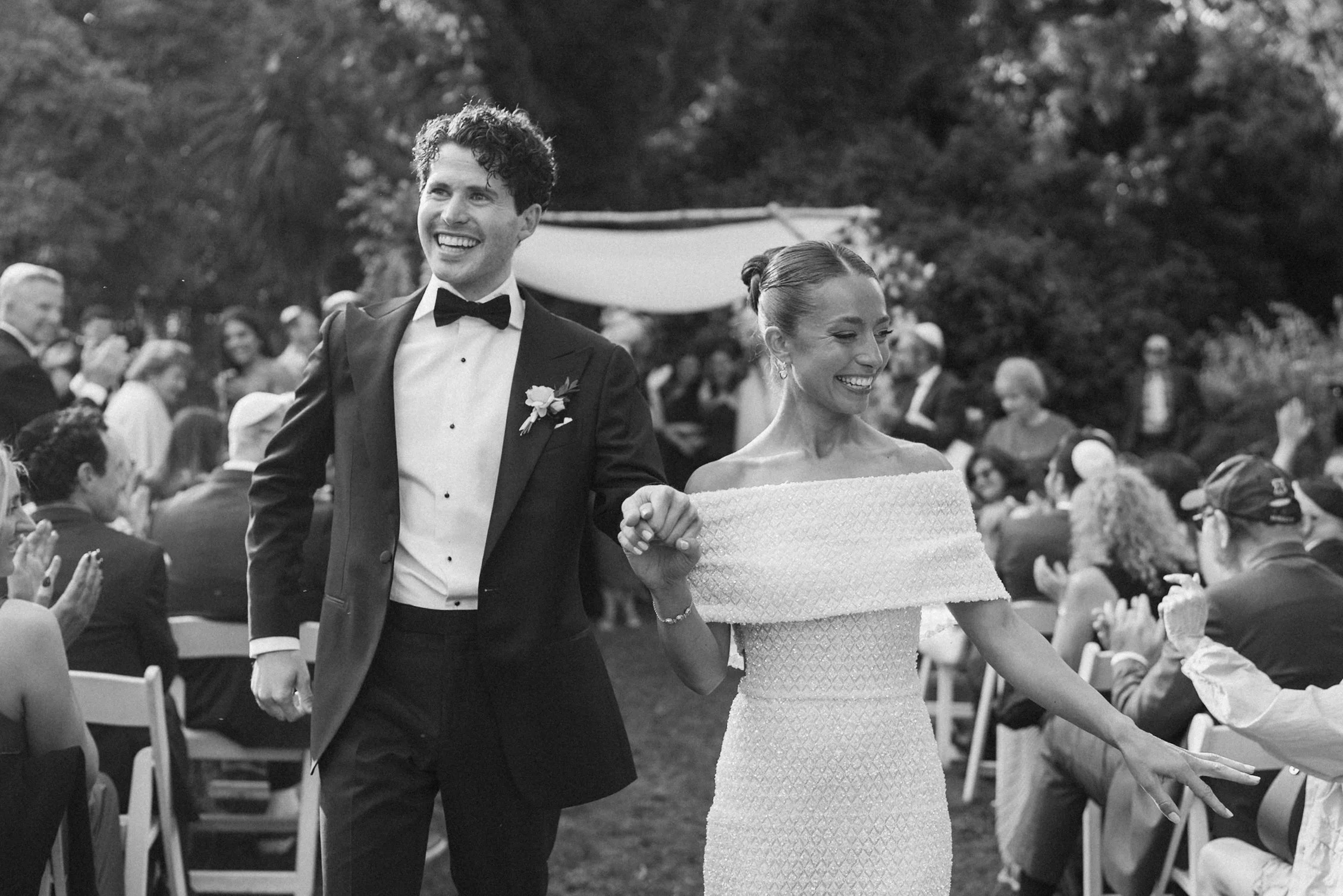 A black-and-white photo of a smiling man and woman at a wedding, walking hand in hand outdoors. The man is dressed in a tuxedo with a bow tie and boutonniere, and the woman in an off-the-shoulder wedding dress. Guests are seated and standing in the b