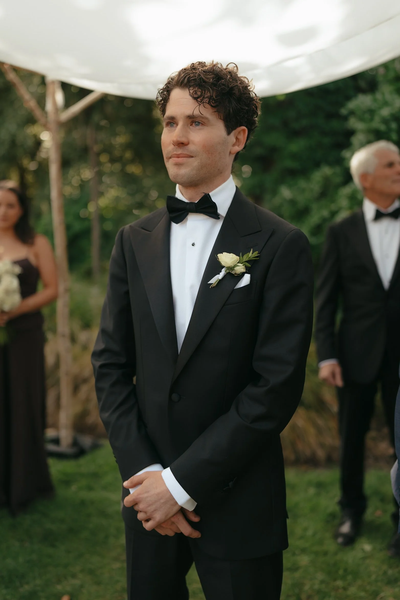 A man in a black tuxedo with a white shirt and black bow tie, wearing a white boutonnière, standing outdoors in front of a green background during a wedding ceremony.