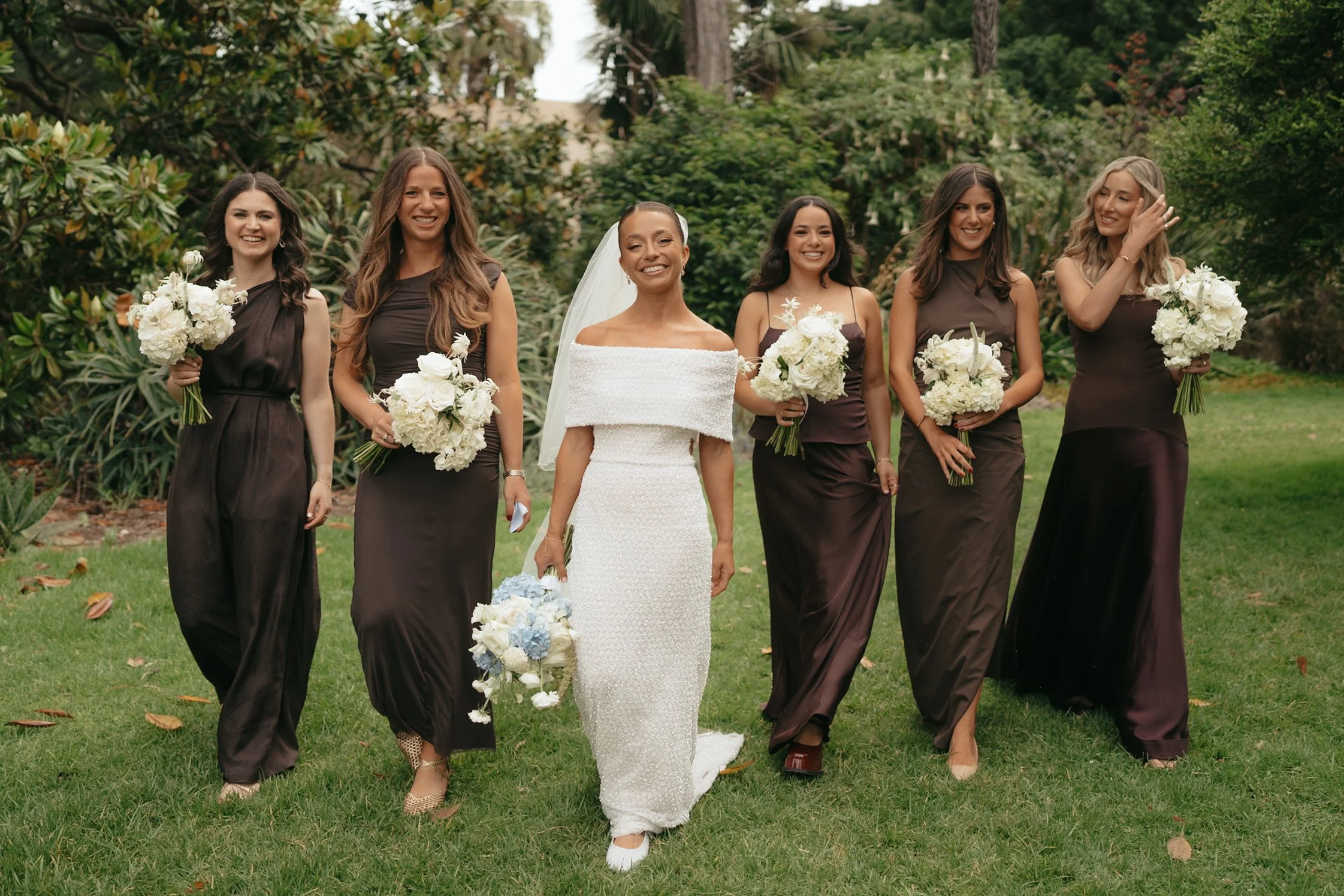 A bride in a white wedding dress walking with five bridesmaids in dark brown dresses holding bouquets of white flowers outdoors in a garden.