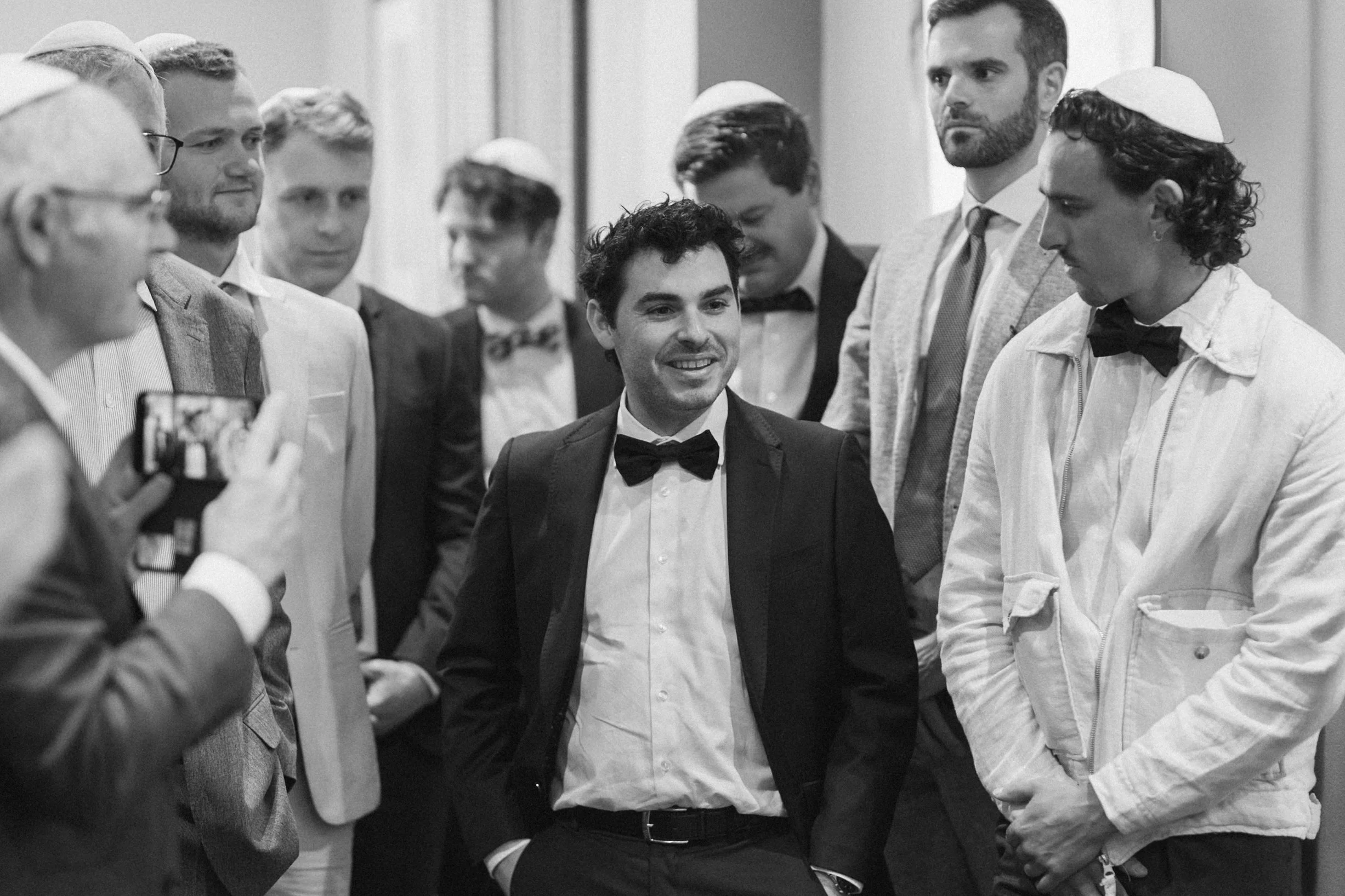 Group of men, some wearing yarmulkes, gathered in a circle, with one man wearing a tuxedo and bowtie smiling at the center, in a formal setting.