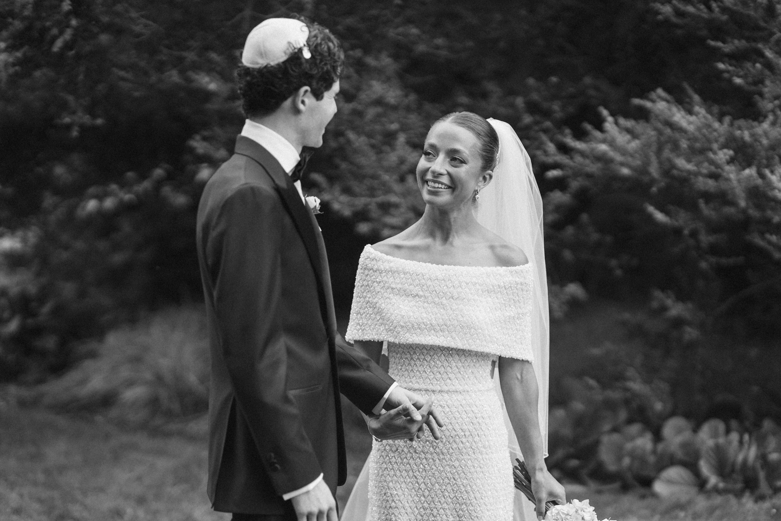 A black and white photo of a bride and groom holding hands during their wedding ceremony outdoors. The groom is dressed in a suit with a boutonniere, and the bride is wearing a textured off-the-shoulder wedding gown with a veil. They are smiling at e