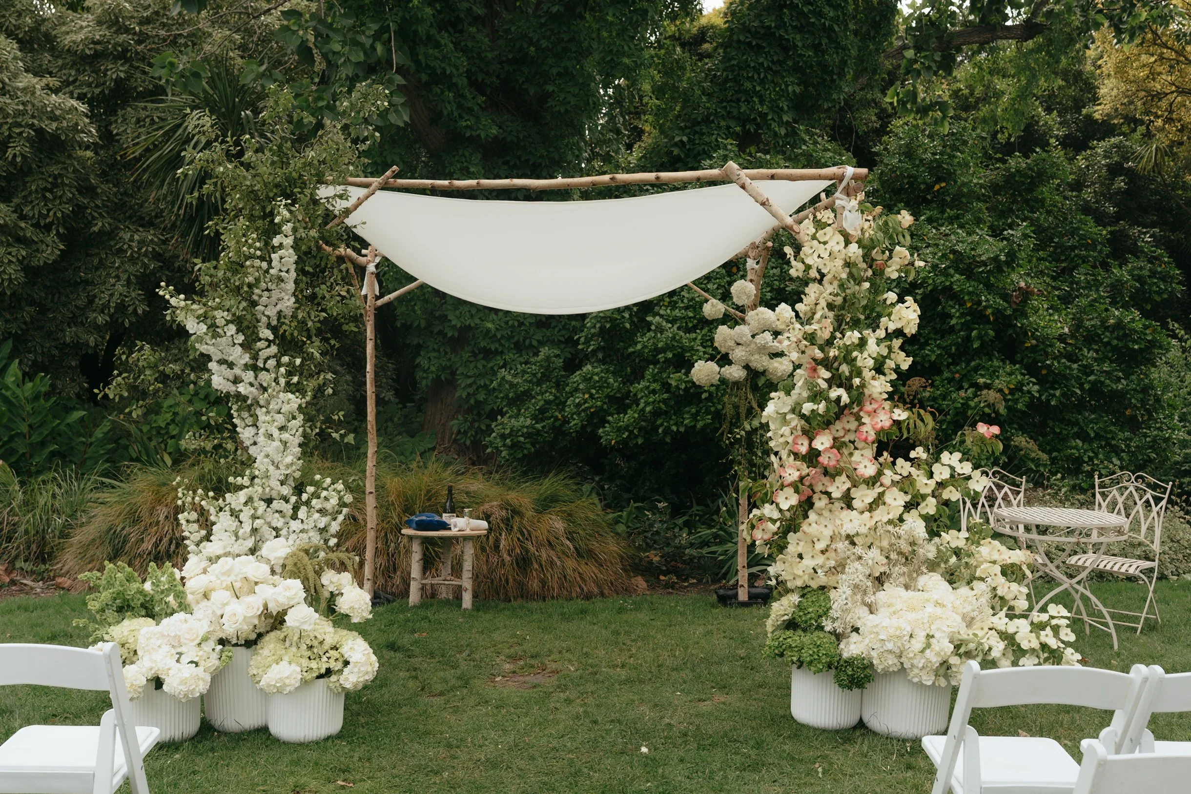Elegant outdoor wedding setup with white chairs, large floral arrangements, and a decorated wooden arch with flowers, set in a lush garden.