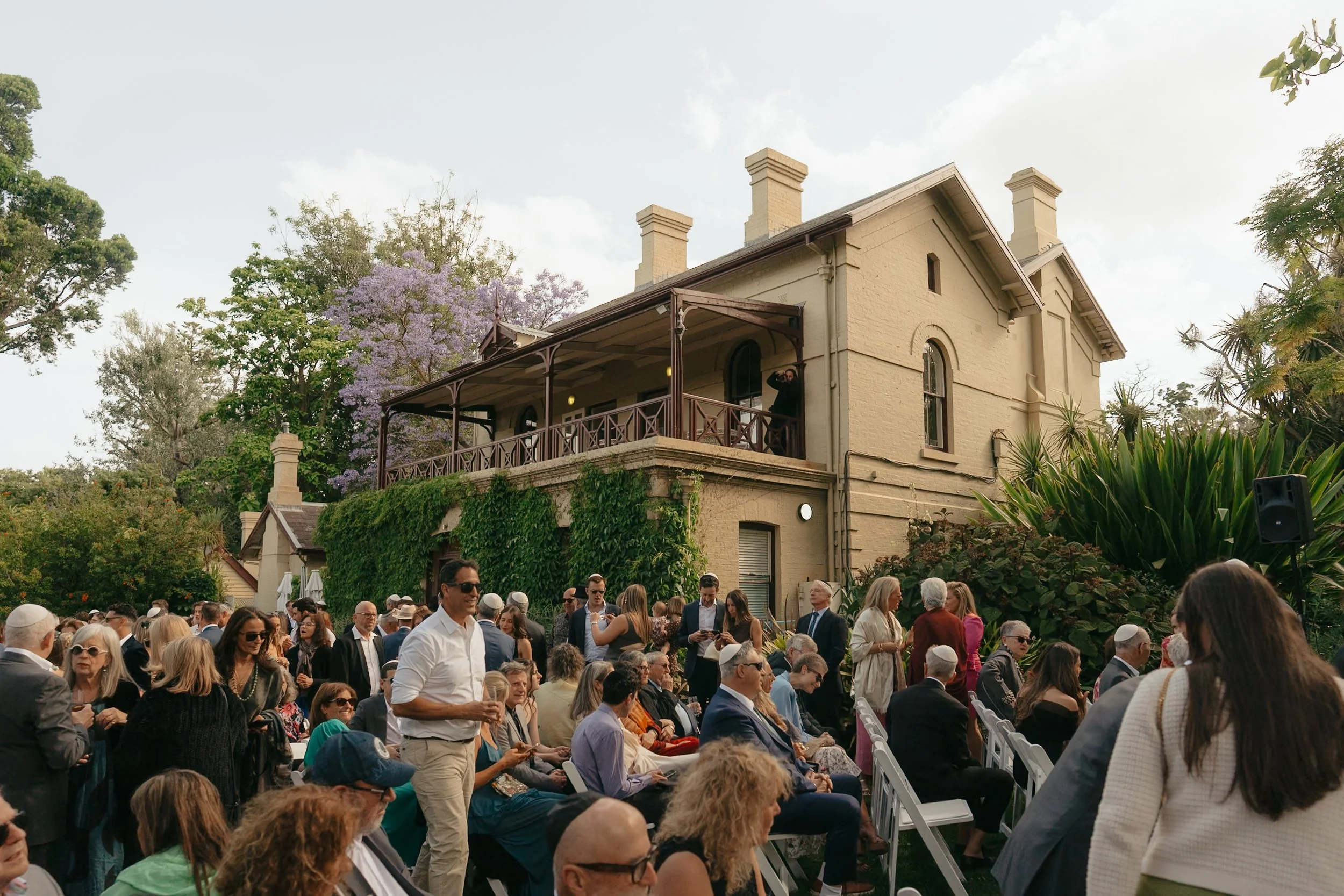 A large group of people gathered outside on a lawn in front of a yellow house with a wooden porch, surrounded by trees, during daytime with partly cloudy skies.