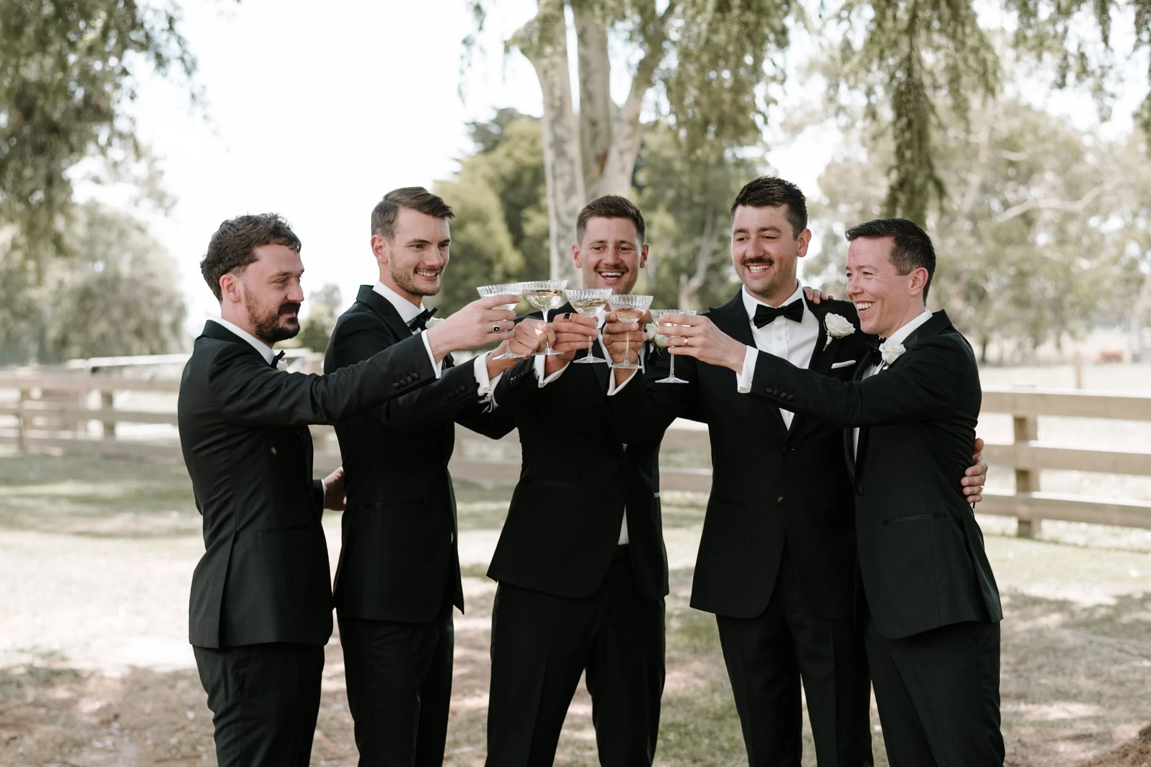 Group of six men in tuxedos clinking glasses outdoors during daytime, smiling and celebrating, with trees and a wooden fence in the background.