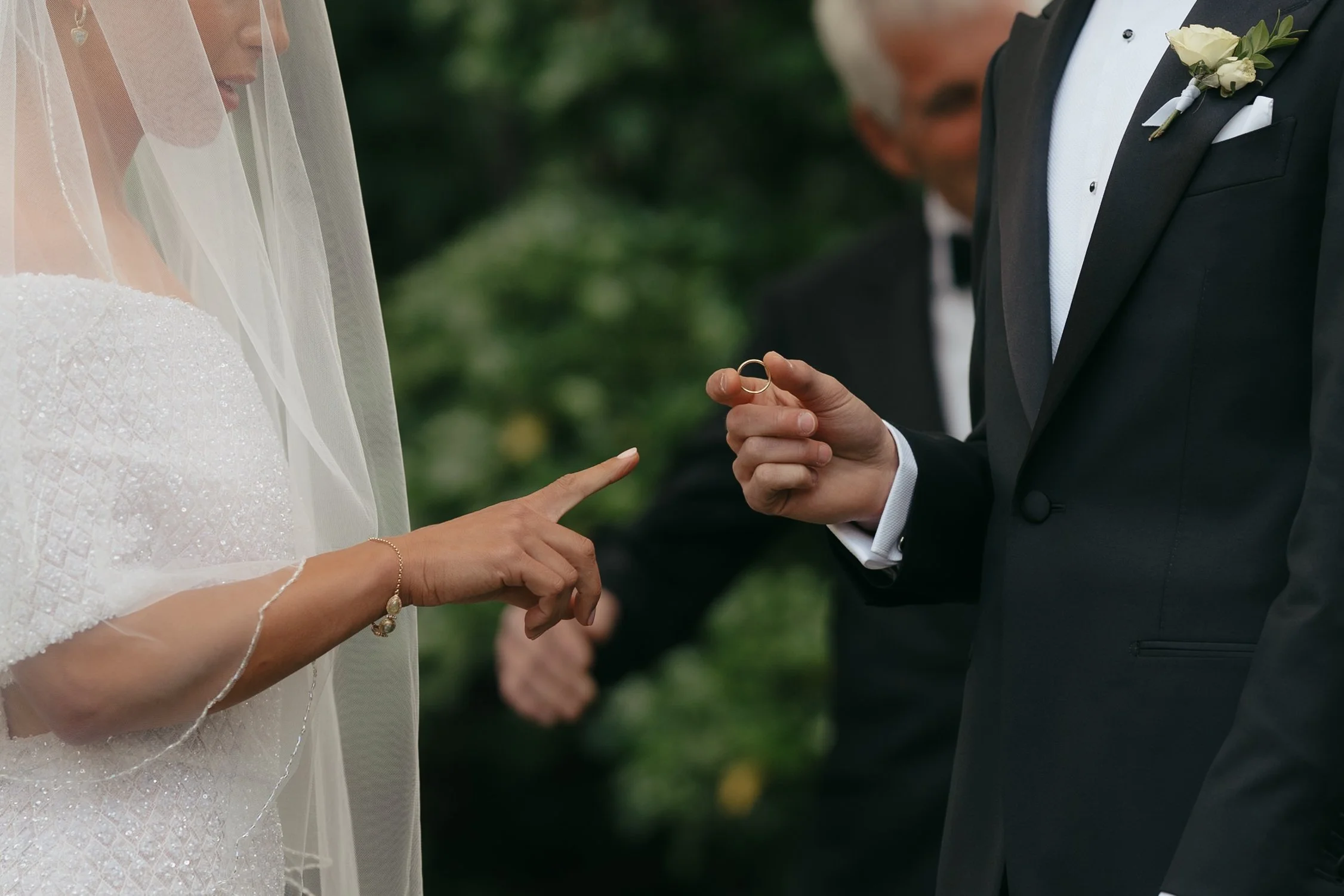 A bride and groom exchanging wedding rings outdoors. The bride is wearing a white wedding dress with a sheer veil, and the groom is in a black tuxedo with a white boutonniere. The groom is holding a wedding band in his hand.