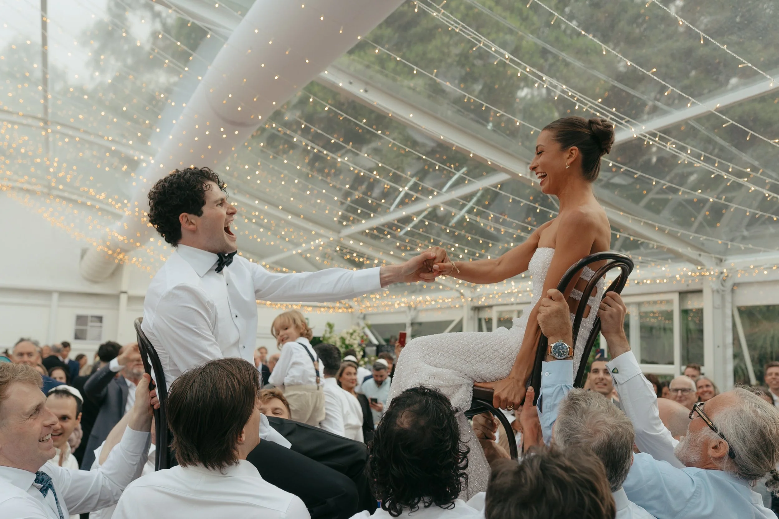 A bride in a white wedding dress is being lifted in a chair by a crowd of people at a wedding reception, with a man holding her hand and smiling. The setting is decorated with string lights overhead, and guests are watching and smiling.