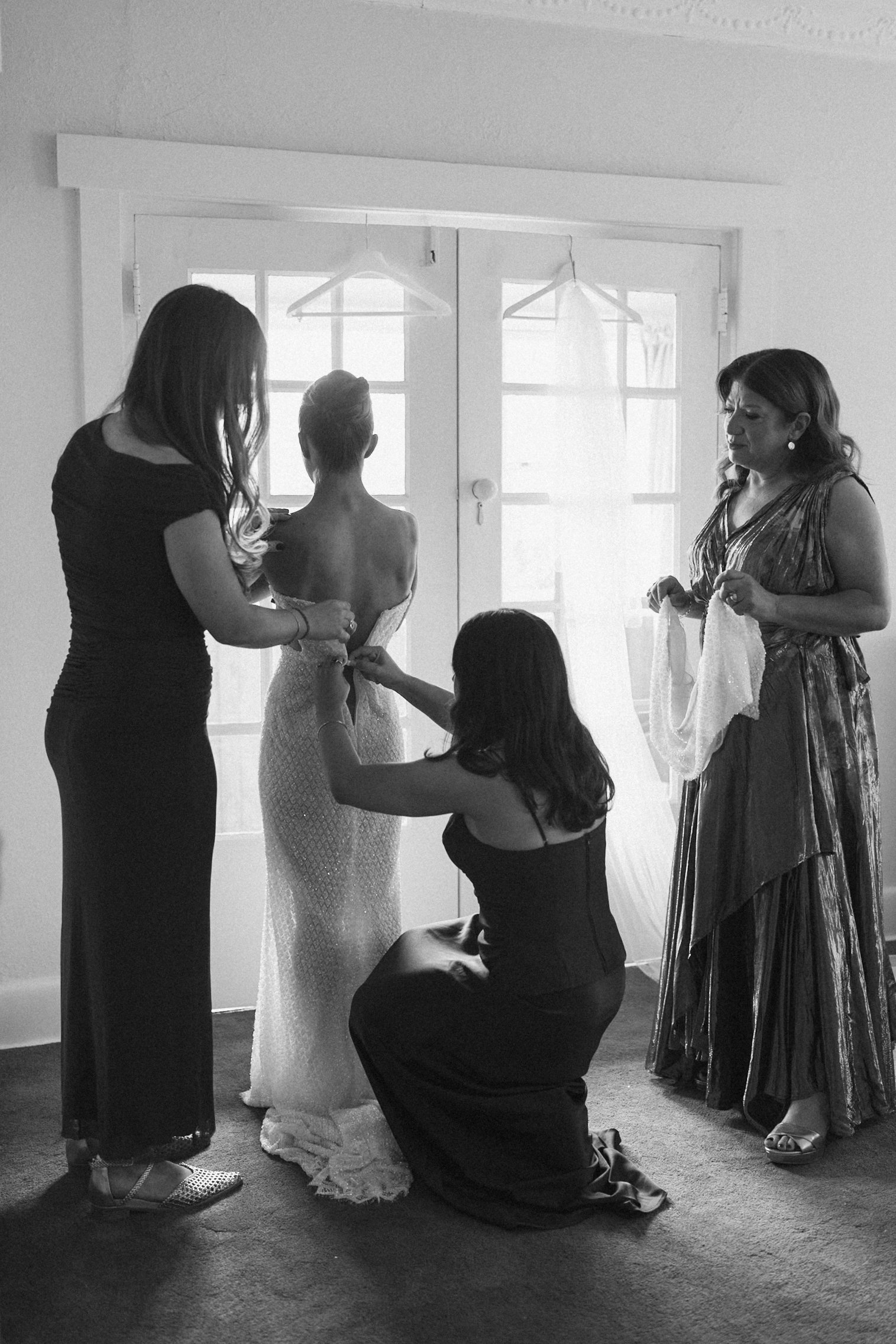 A bride getting ready for her wedding, assisted by her bridesmaids in a room with large windows. The woman on the right holds a towel, and others help with the bride's dress, which is strapless and fitted.