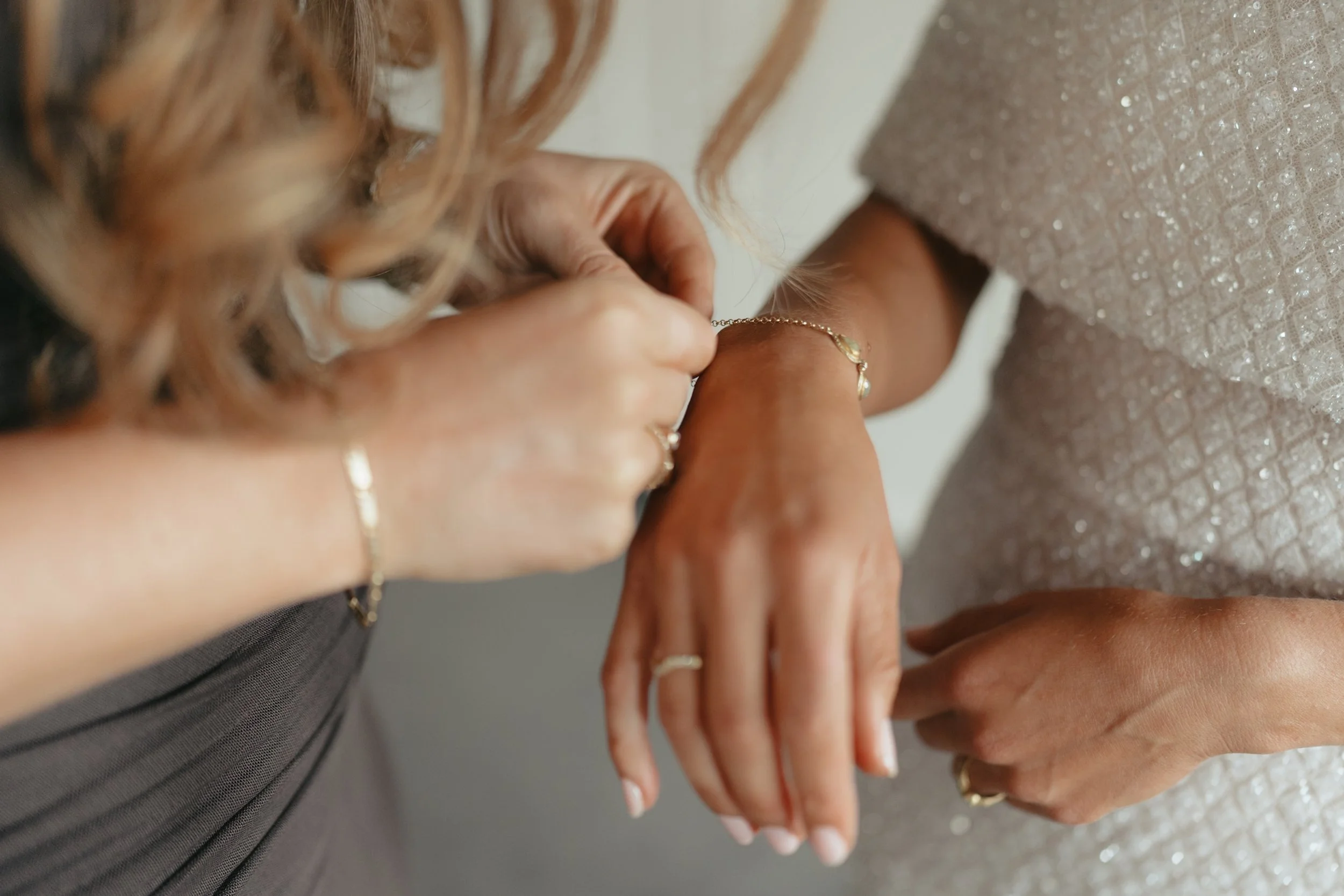 Close-up of two women holding hands, one wearing a gray dress with sparkles and the other wearing jewelry, possibly at a wedding or special occasion.