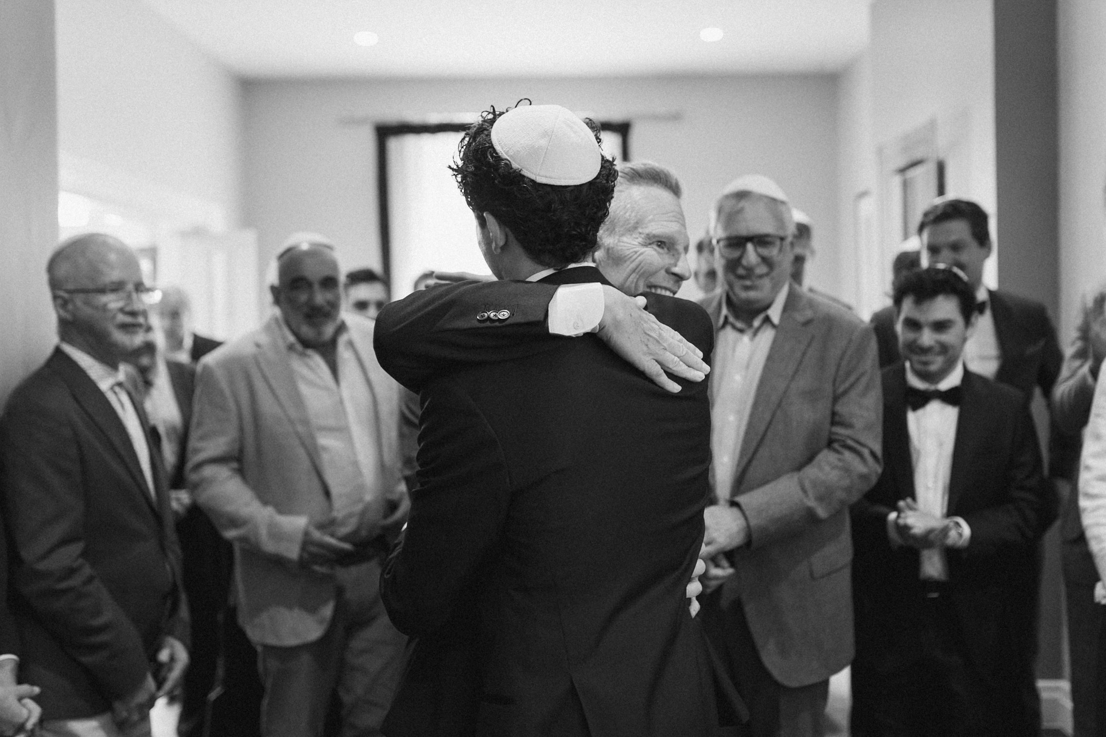 A group of men in suits and ties gathered indoors, with two men embracing in the center, one wearing a yarmulke, amid smiling and attentive onlookers.