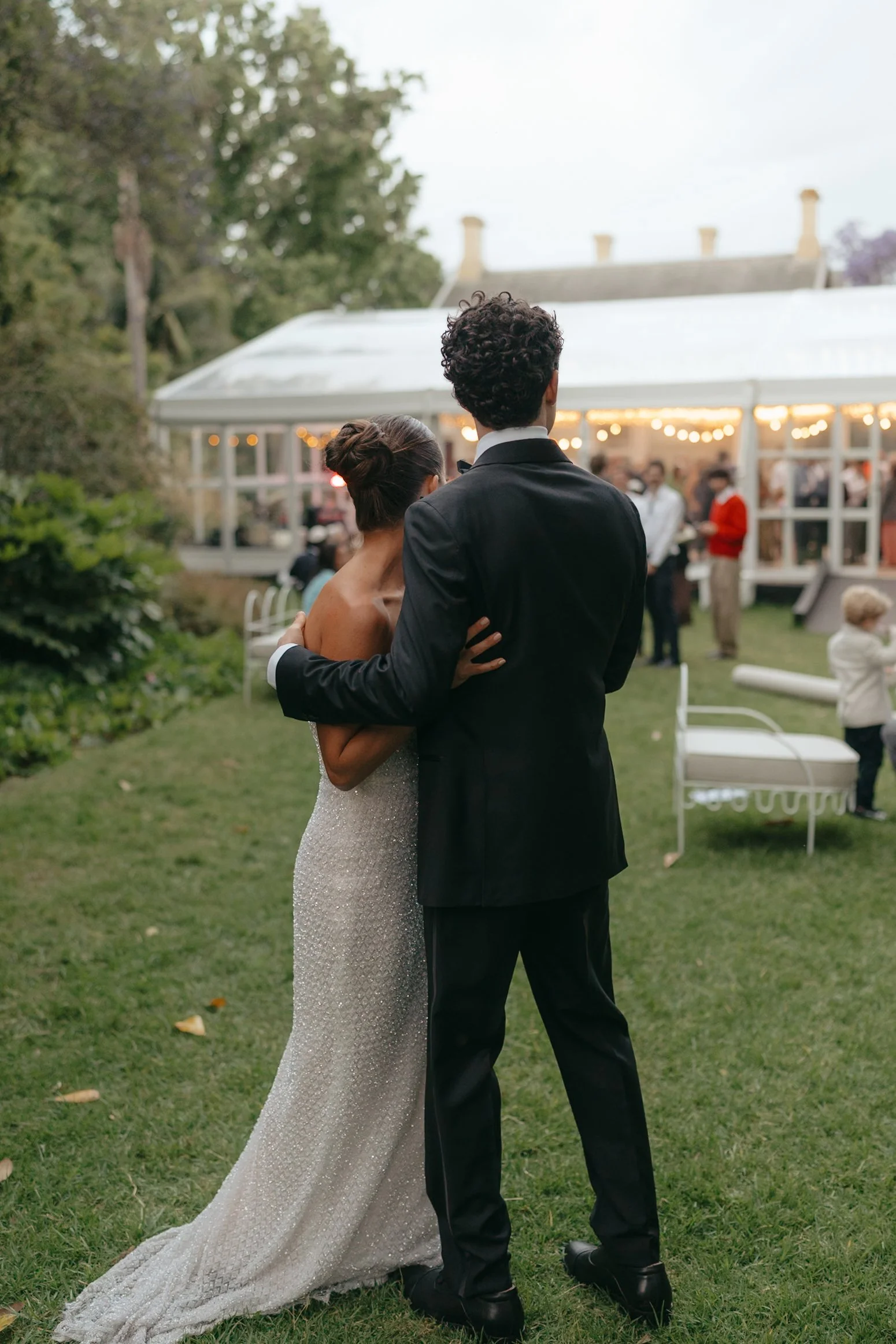 A bride and groom dressed in formal wedding attire, standing outside at a wedding reception under a tent, with guests in the background.