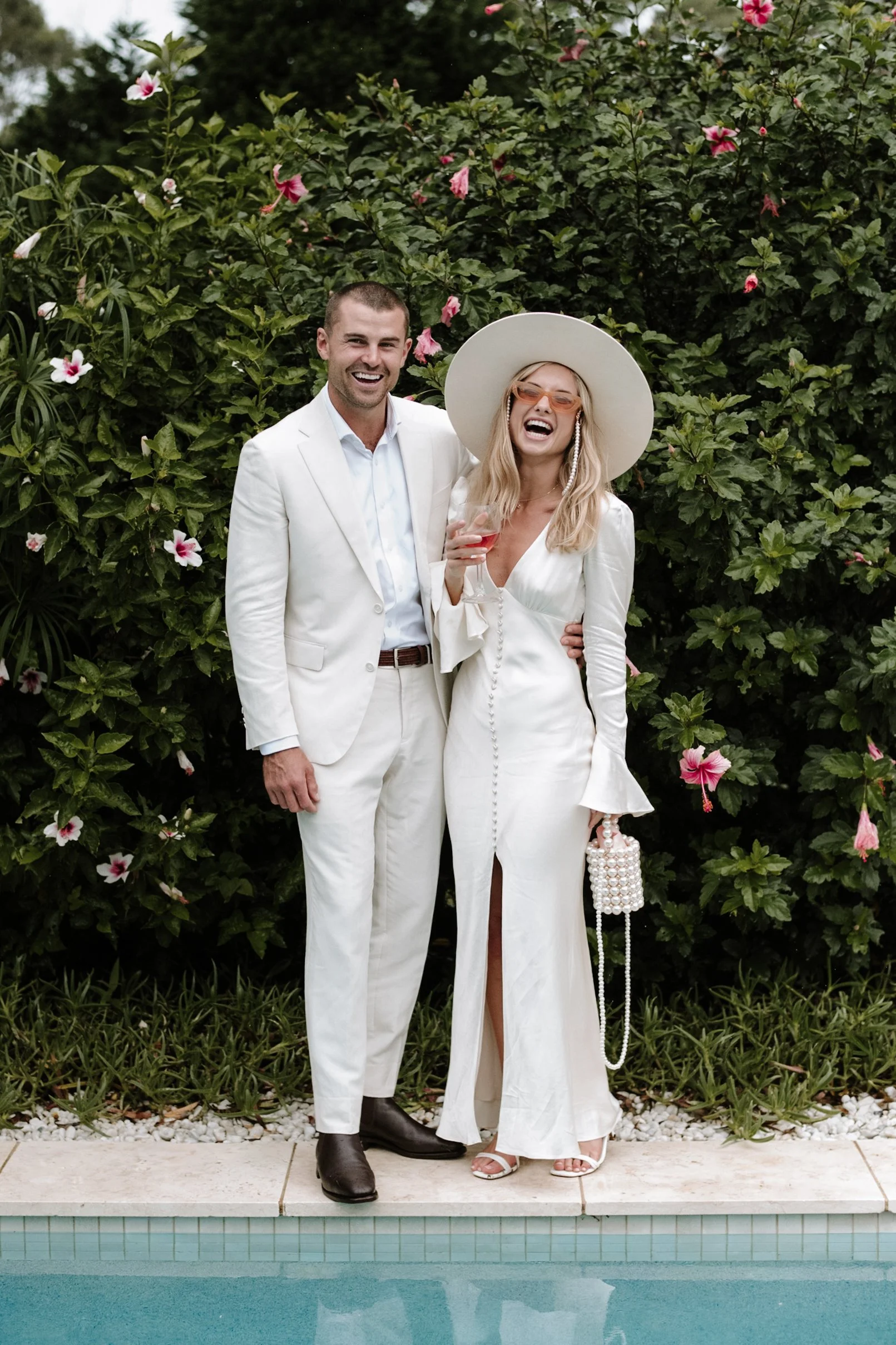 A man and woman dressed in white, standing by a pool, smiling and laughing. The woman is wearing a large white hat, sunglasses, and holding a glass of rosé wine. The background features lush green bushes with pink and white flowers.