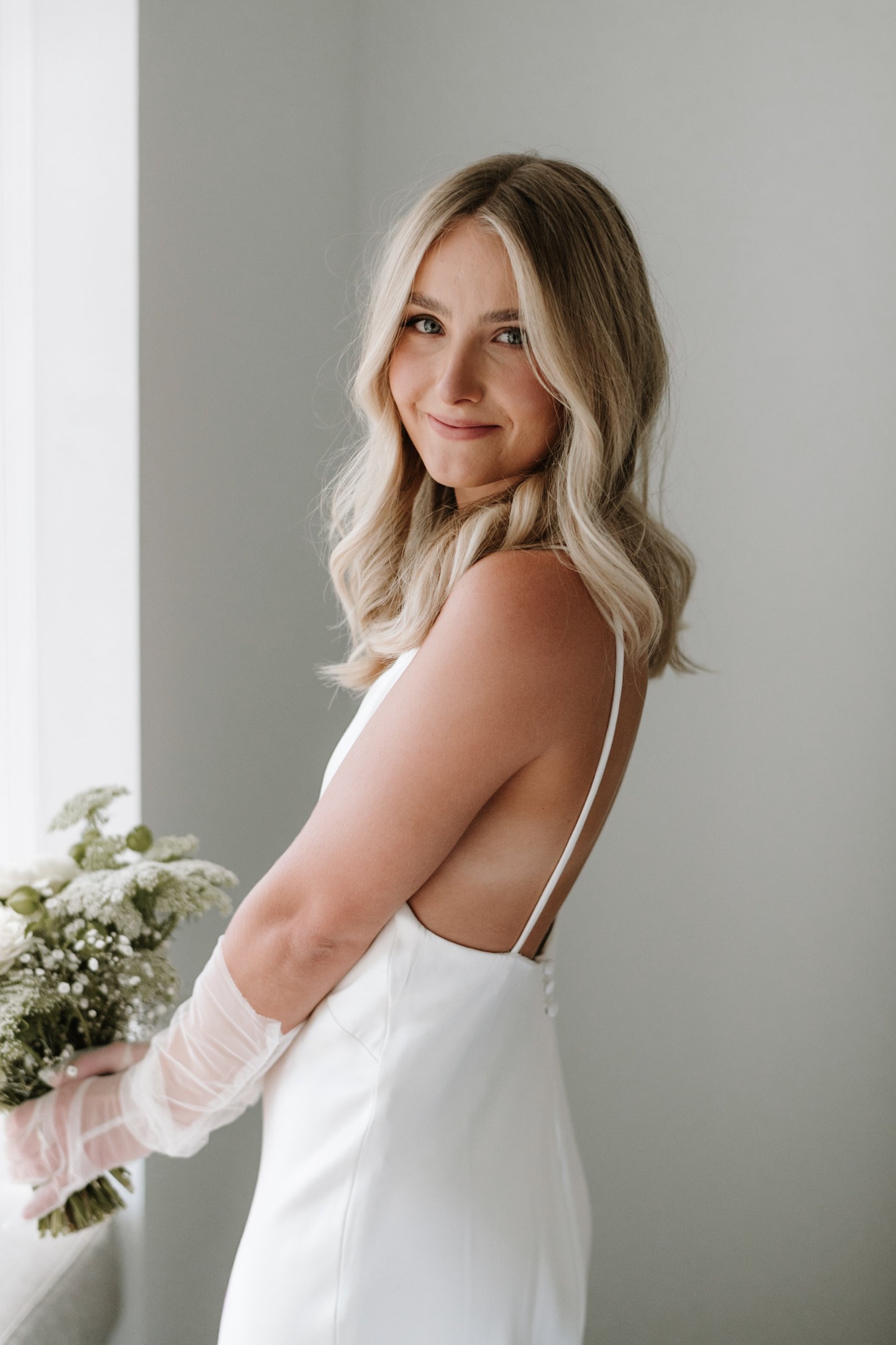 A young woman with blonde hair styled in soft waves, smiling softly while holding a bouquet of white flowers. She is wearing a white dress with thin straps, standing against a plain, light-colored wall.
