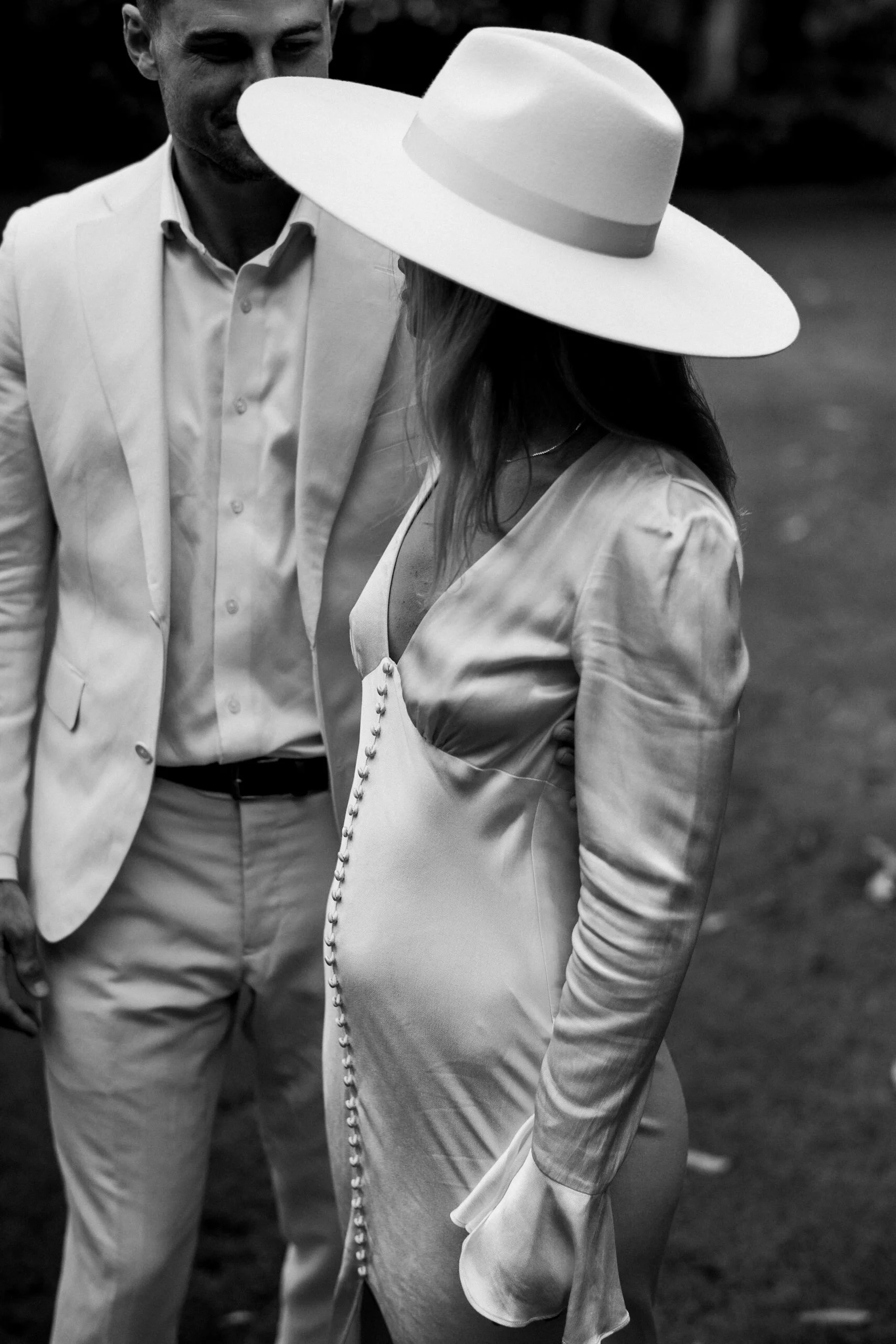 A man and woman dressed in formal attire, with the woman wearing a large brimmed hat, stand close together outdoors.