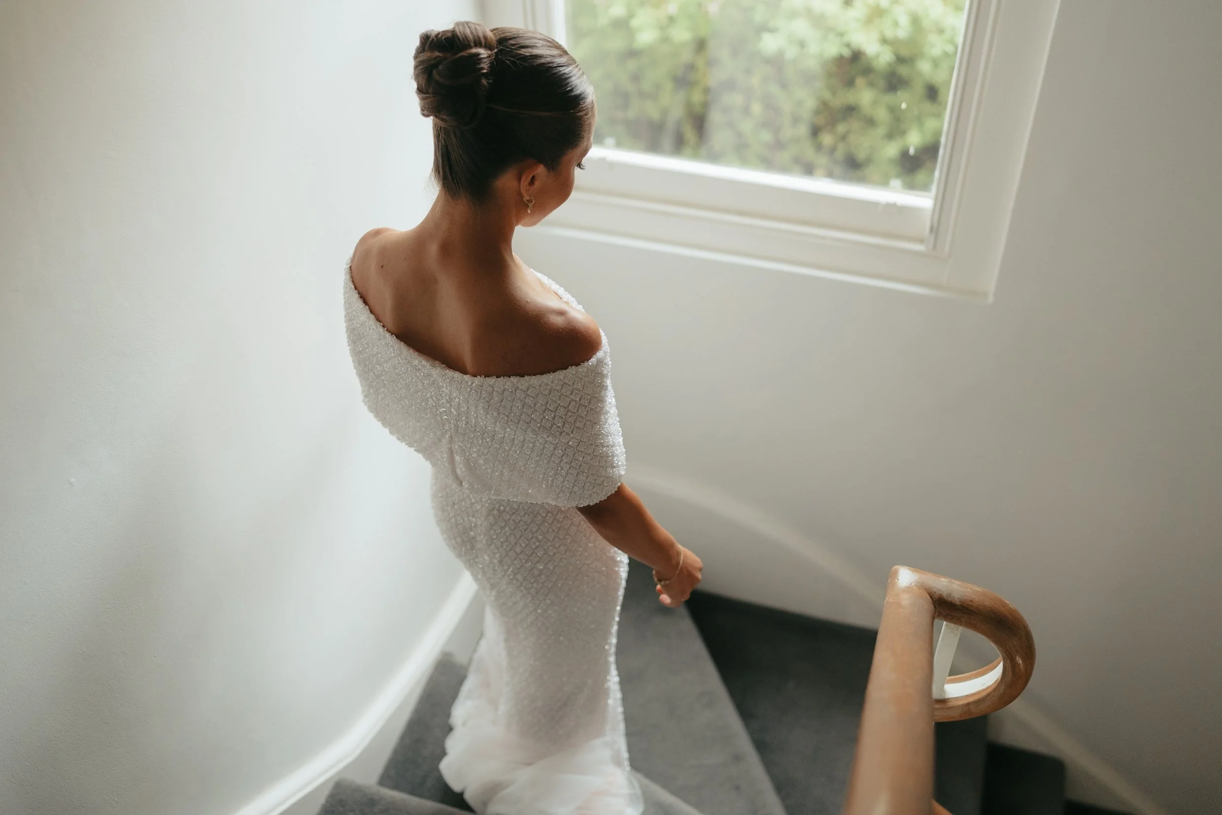 A woman with dark hair styled in a high bun, wearing a white off-shoulder dress with a textured pattern, is looking out a window. She is standing indoors near a staircase with wooden handrails, in a well-lit room with white walls and a large window s