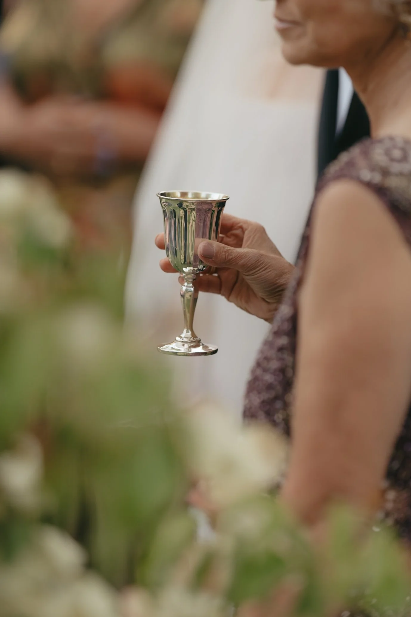 A woman in a floral dress holding a silver goblet, with a blurred background of greenery and other people at a formal outdoor event.
