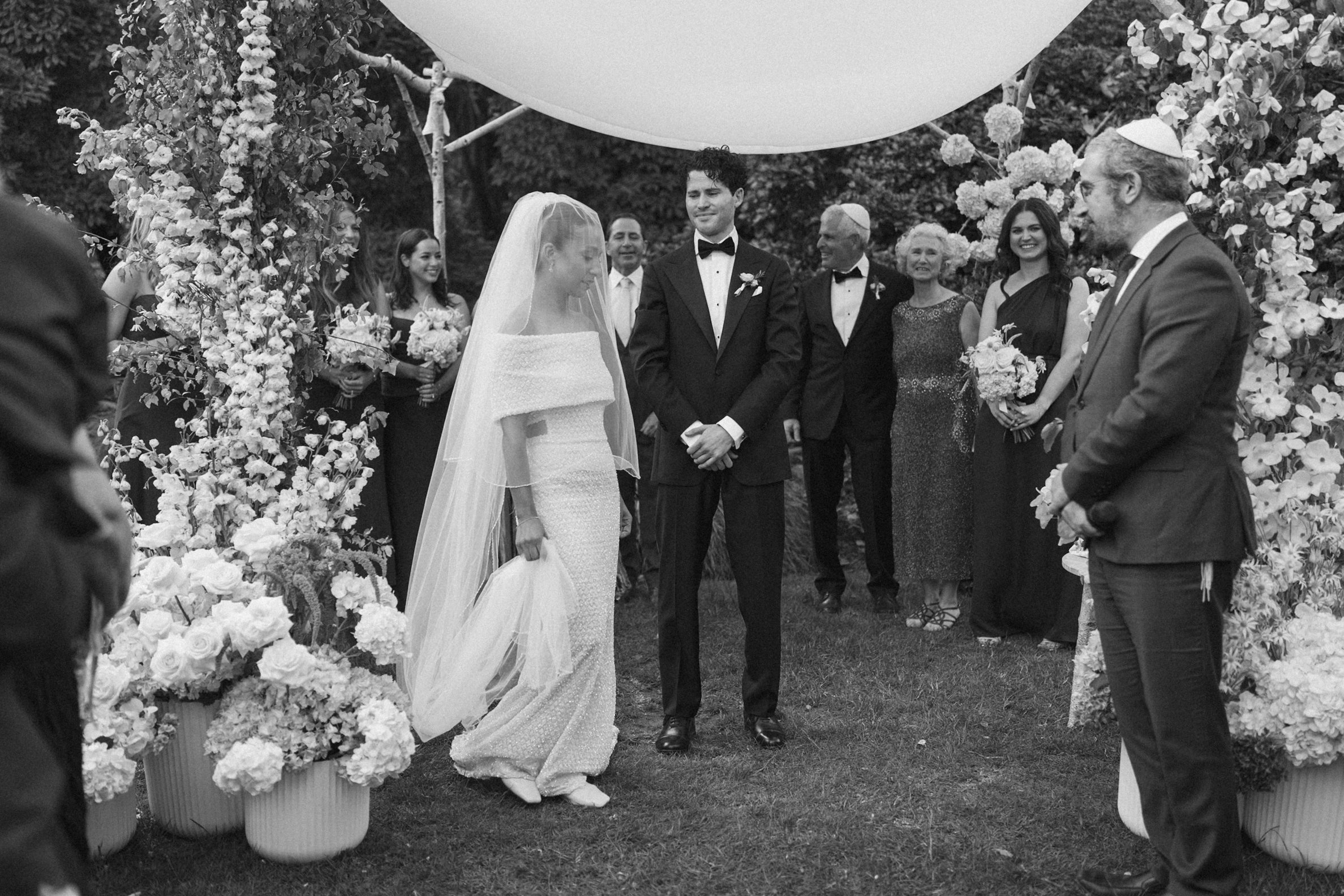 A black and white photo of a wedding ceremony outdoors, with a bride and groom standing in front of an officiant. The bride is wearing a traditional dress and veil, and multiple guests, dressed formally, are standing around them, smiling. The scene i