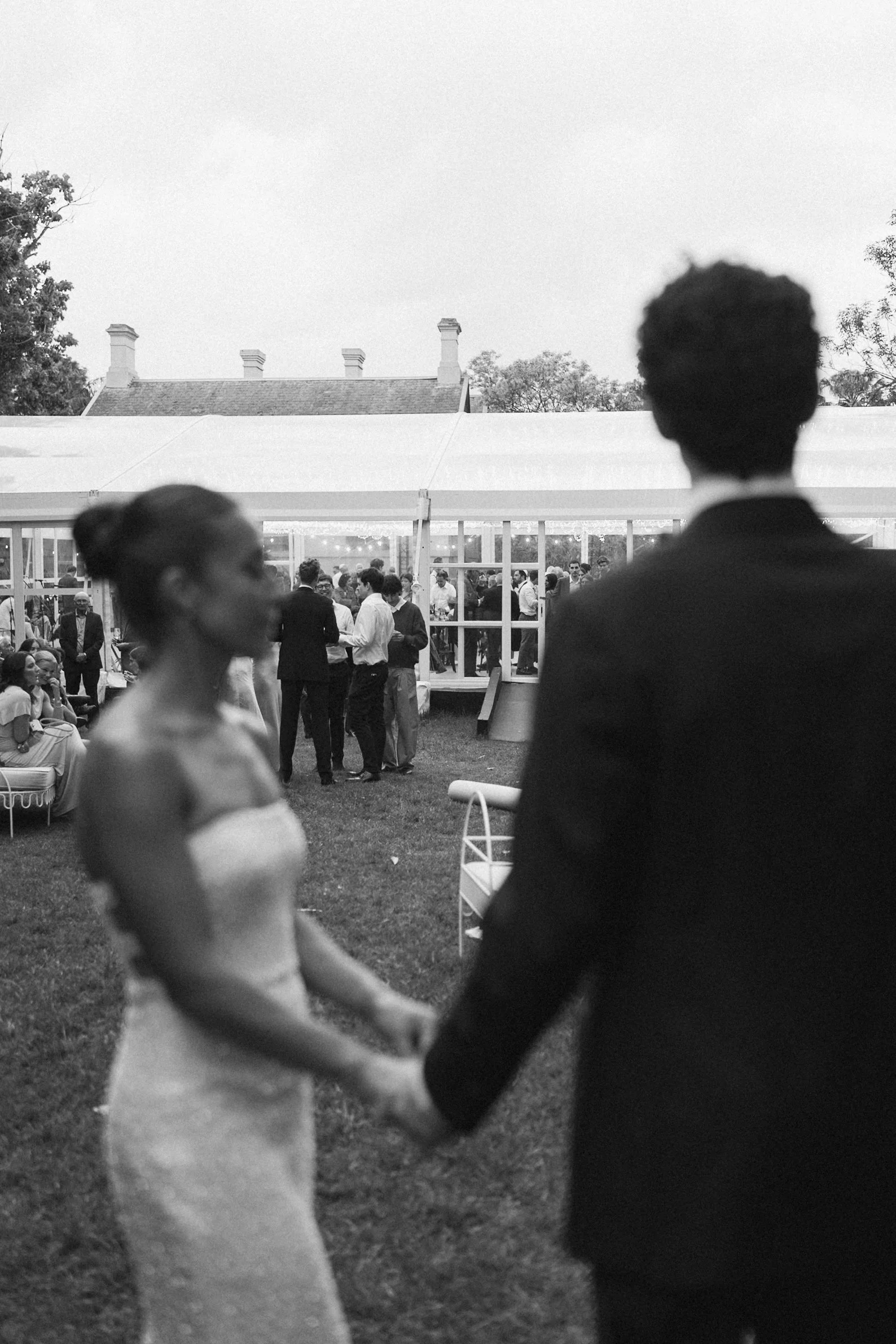Two people holding hands and dancing outdoors at a gathering, with many guests in the background near a large tent, in front of a house and trees.