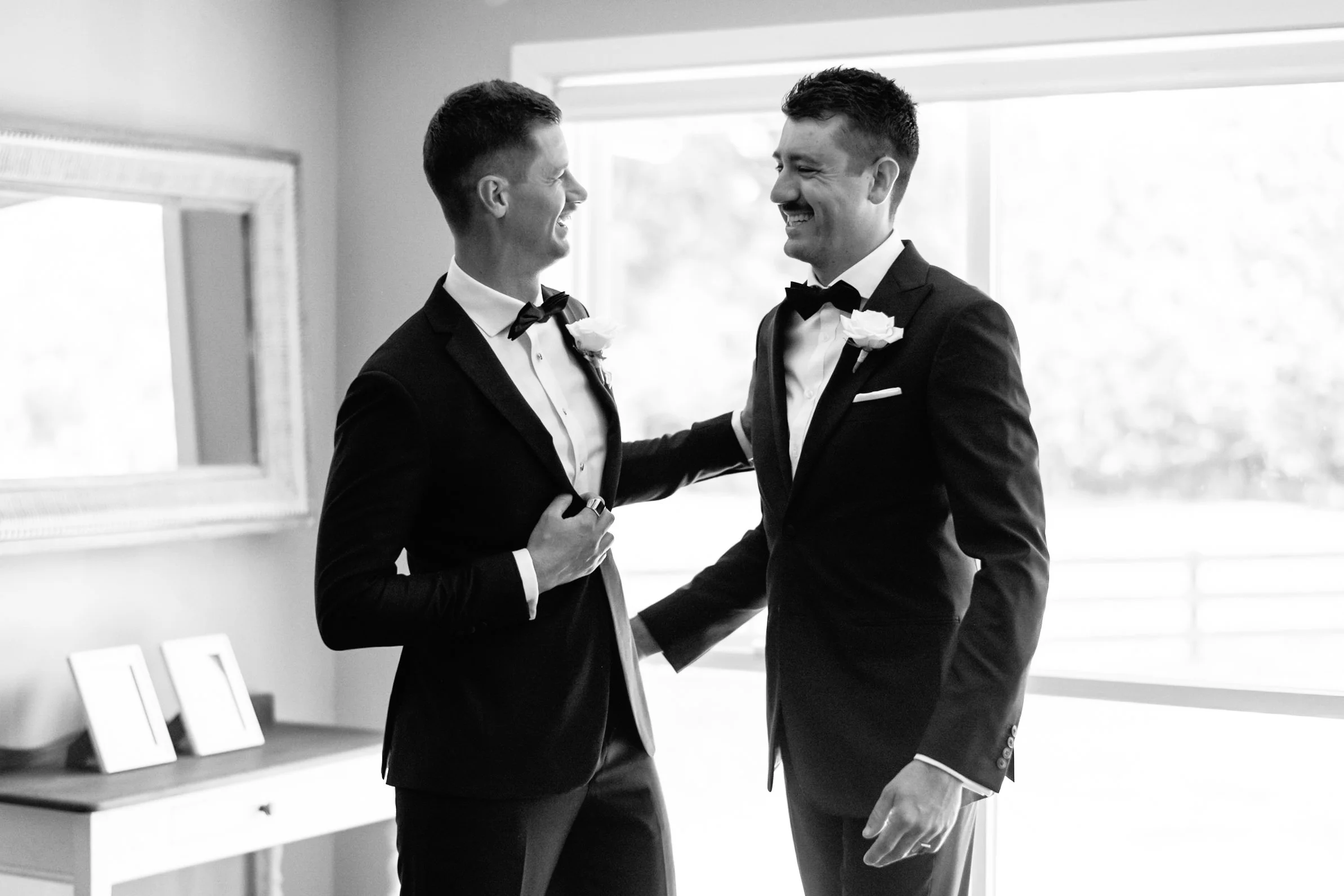 Two men in tuxedos smiling and holding hands in a well-lit room, possibly during a wedding or special event.