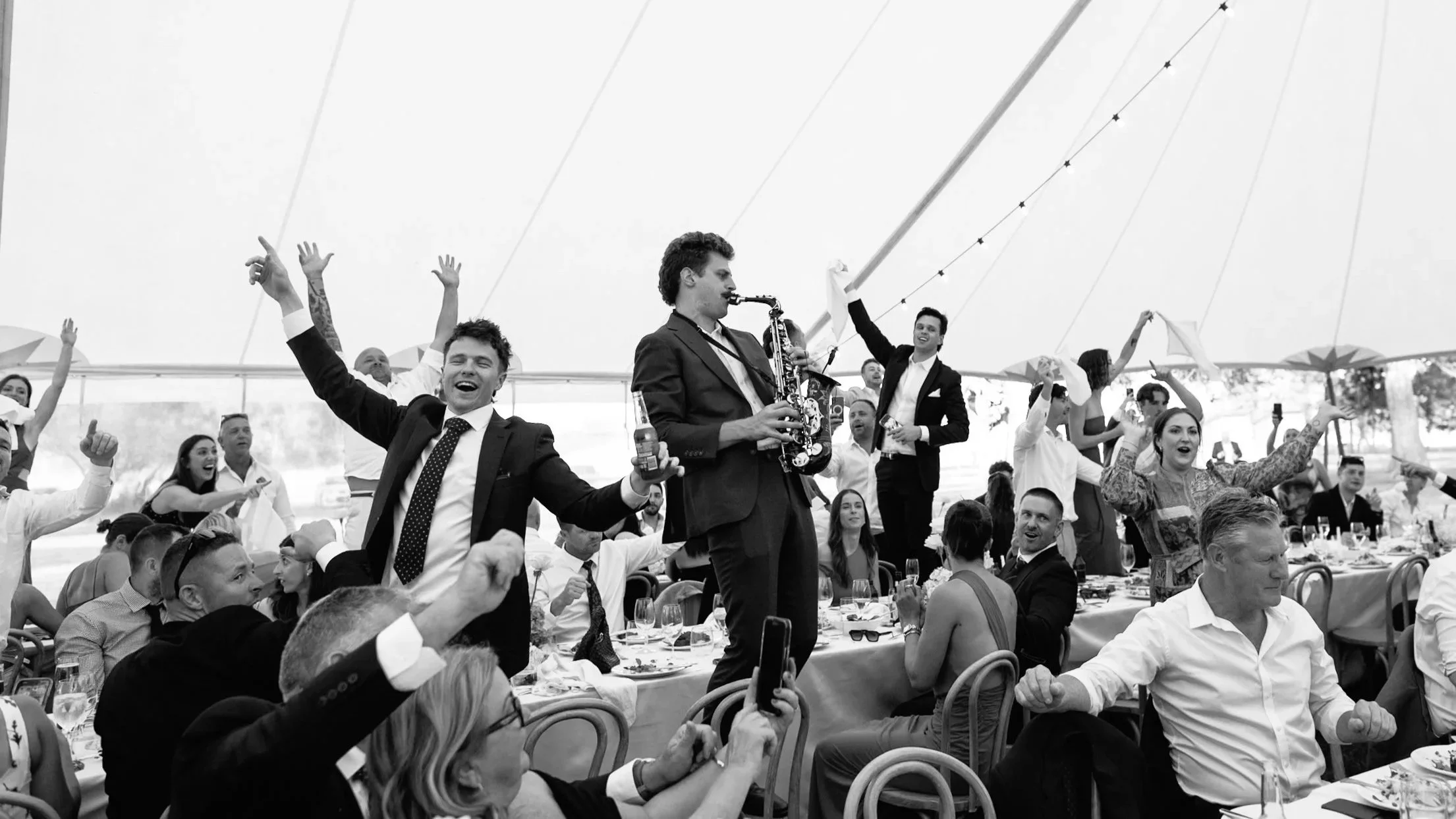 A black and white photo of a lively gathering with many people seated at tables, some raising their hands or glasses, while a musician in a suit plays the saxophone on a raised platform.